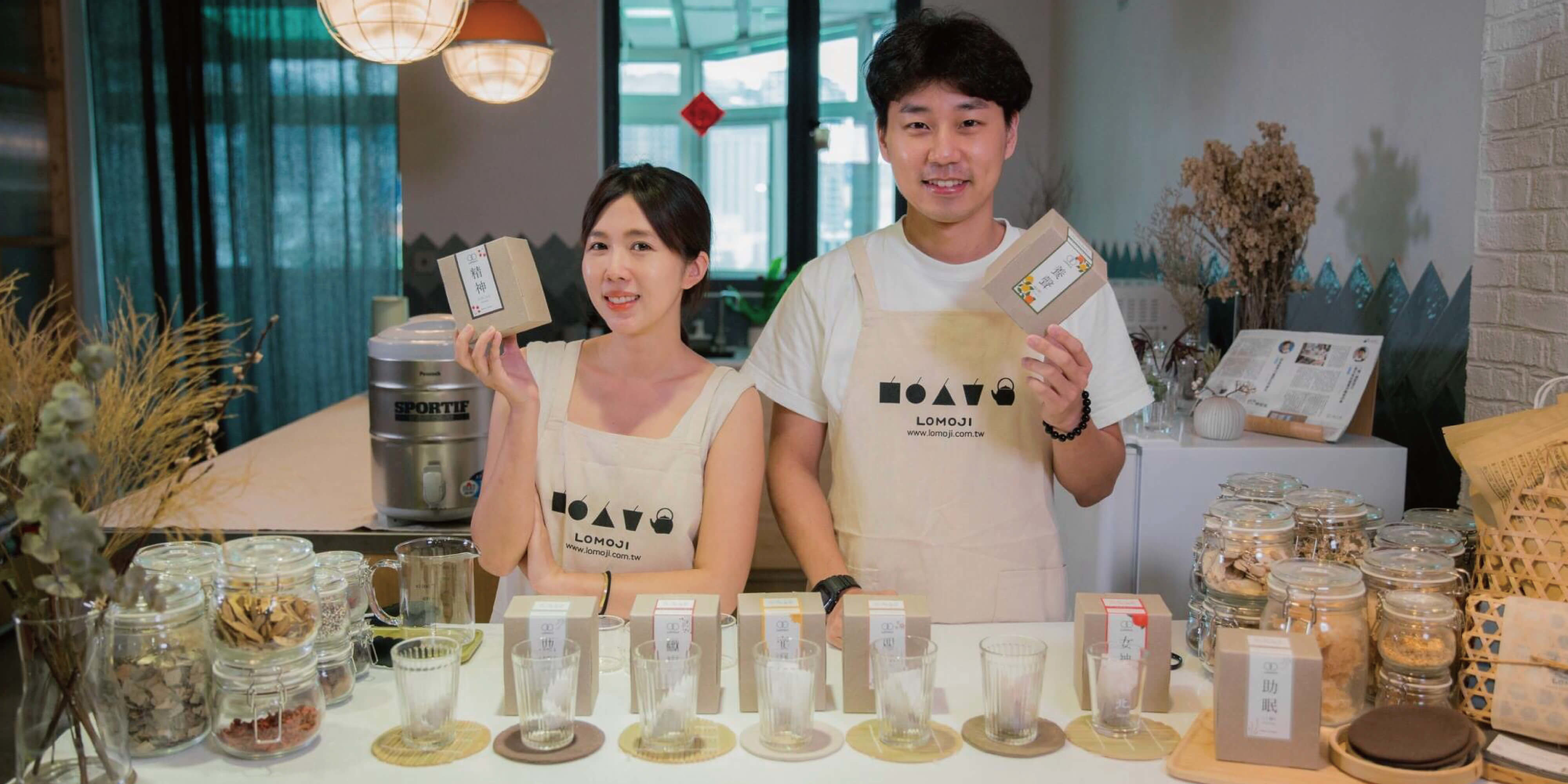 樂木集創辦人Hung與April拿著產品，桌上擺有樂木集的漢方茶與玻璃杯