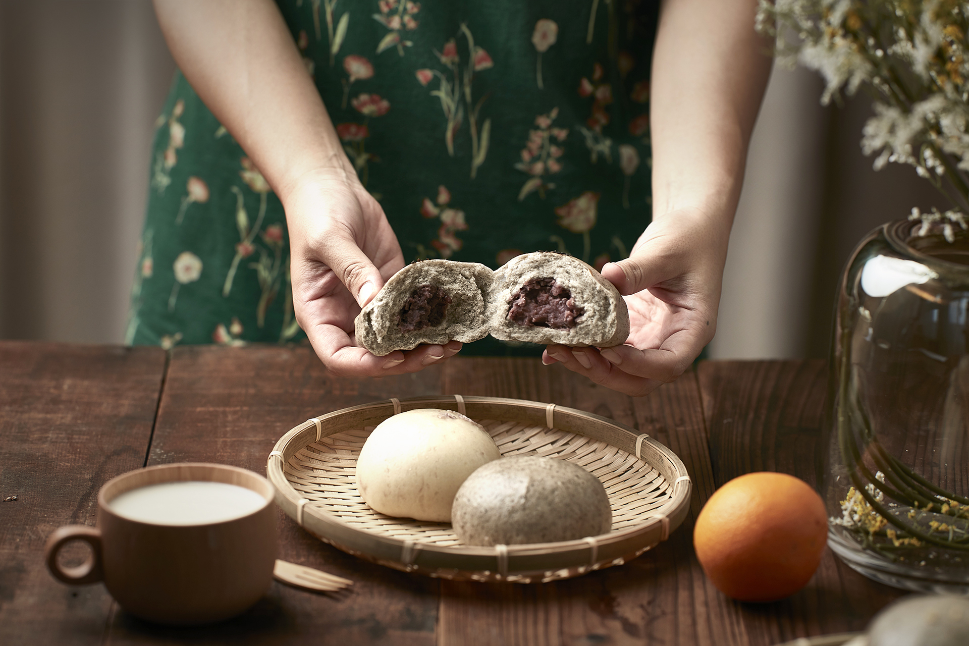 桌面上的竹籃裡放著兩個包子，一位女性站在桌子後方，雙手正剝開一只紅豆包。