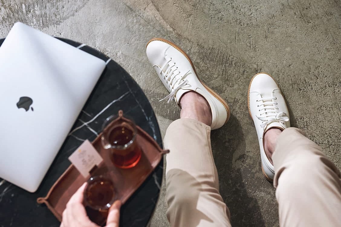 皮革休閒鞋 俯視身穿卡奇休閒褲的模特搭配白色皮革休閒鞋坐在擺有筆記型電腦的桌子旁