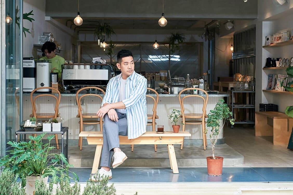 皮革休閒鞋 穿著清爽藍白休閒襯衫搭配白色皮革休閒鞋的模特坐在咖啡廳外的長木椅上望向左方