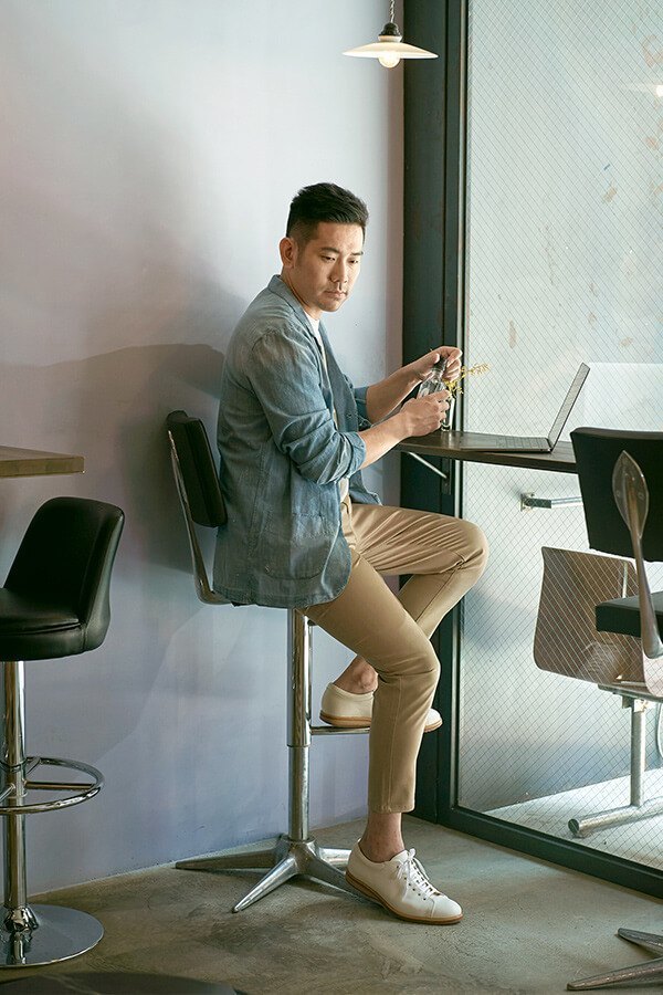皮革休閒鞋 穿著牛仔襯衫卡其褲搭配白色皮革休閒鞋的模特坐在窗邊的吧檯使用筆記型電腦