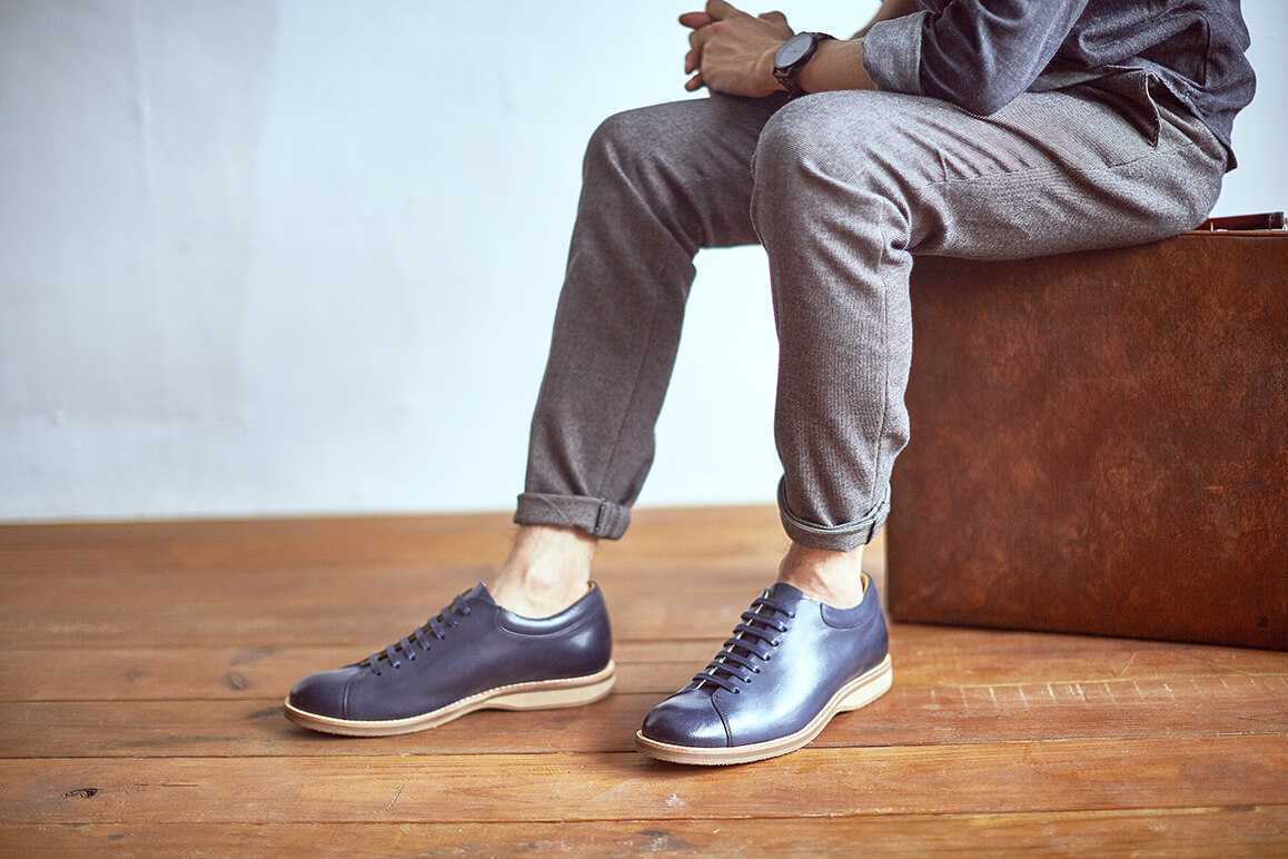 穿著闇夜藍色皮革休閒鞋搭配兔灰色休閒西裝褲的模特兒坐在皮革手提箱上