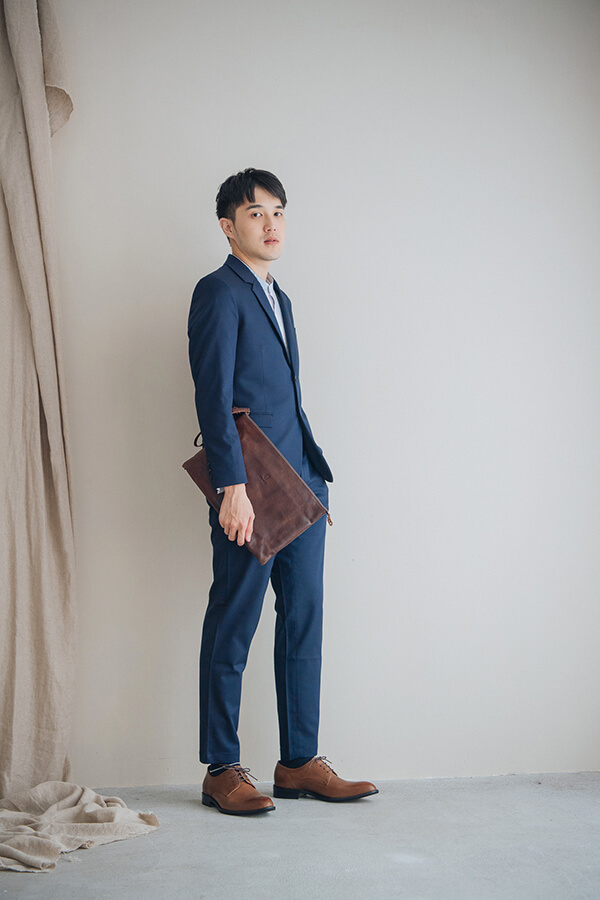 穿著一套湛藍色西裝的模特兒站在淡米色的牆面前右手拿著咖啡色皮革文件包搭配咖啡色德比鞋紳士鞋