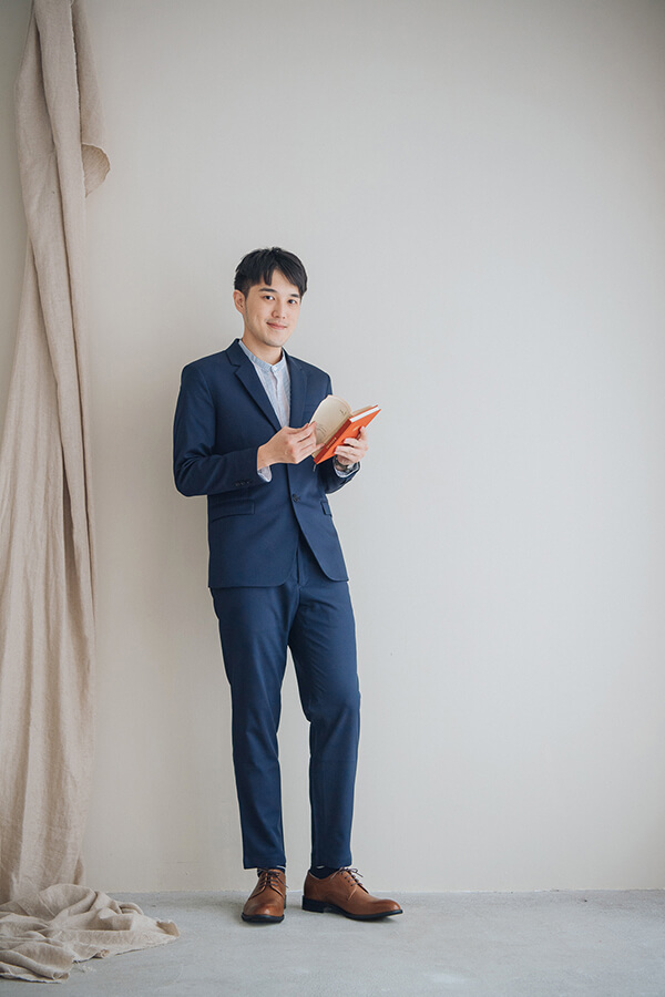 穿著一套湛藍色西裝的模特兒背靠在淡米色的牆面前雙手拿著橘色的書本腳上穿著林果良品的咖啡色德比鞋紳士鞋