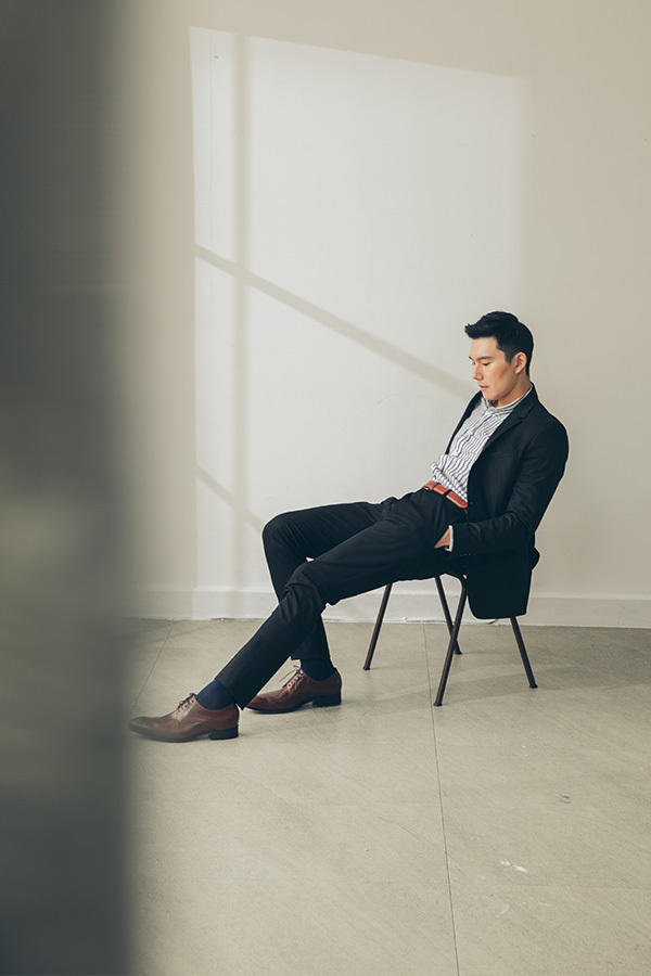 椅子上的模特穿著正式黑色商務西裝搭配可可咖啡色經典流線德比鞋