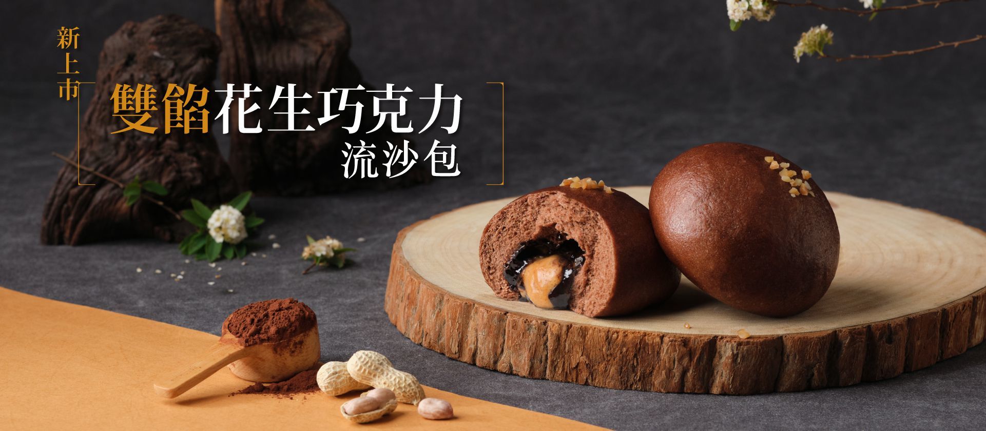 兩顆深咖啡色的花生巧克力流沙包陳列在木質圓盤上，其中一顆被剝開，流出花生與巧克力的內餡。畫面前方有一匙可可粉以及剝開的帶殼花生。