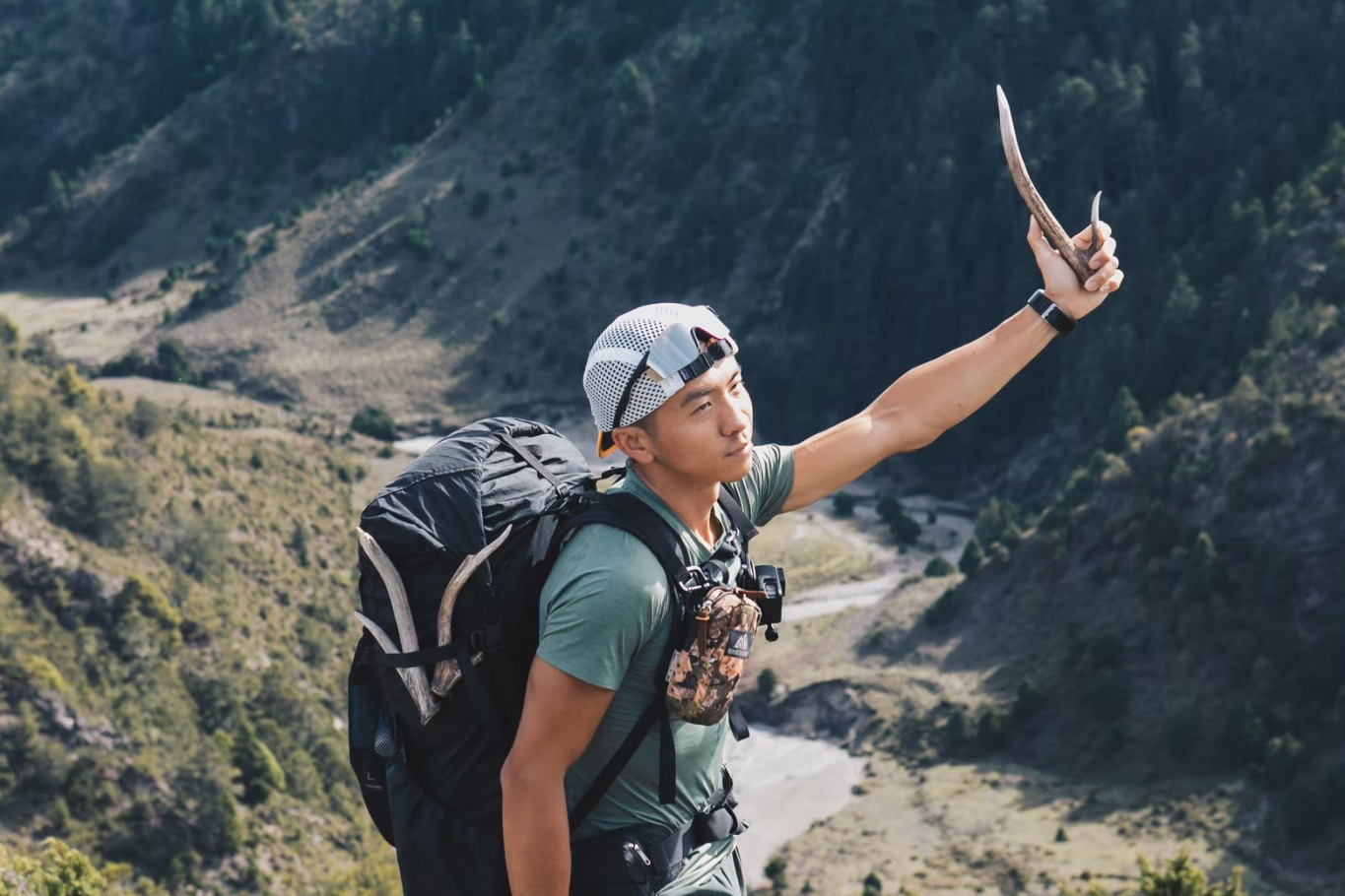 張岱偉身穿綠色衣服，背著黑色後背包在山中，左手拿著鹿角。