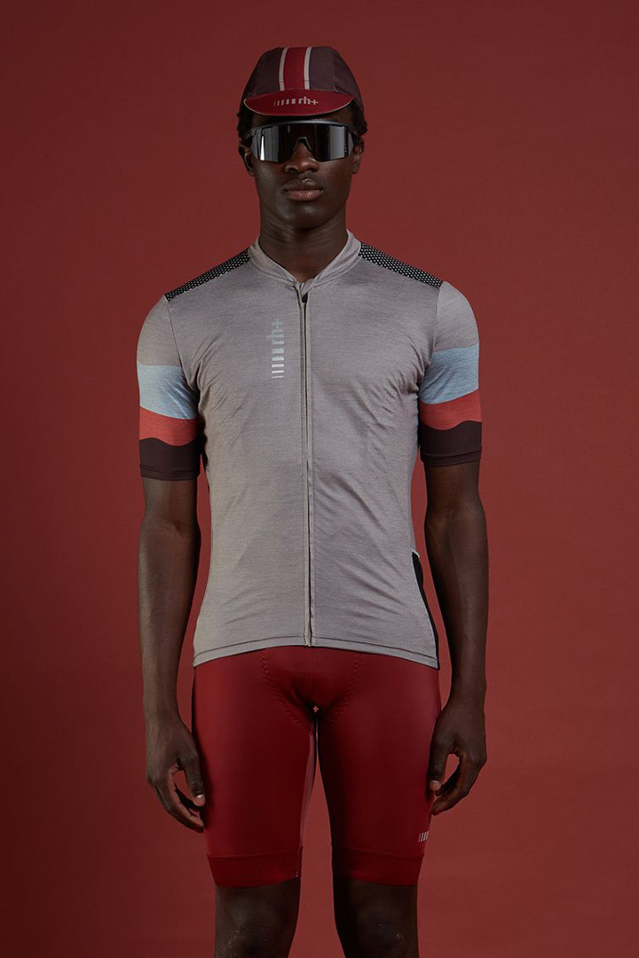專業模特兒在背景紅色的地方身穿灰色TOUS TERRAIN系列專業自行車衣加紅色PRIME系列專業自行車褲,頭上戴著自行車小帽和戴著運動眼鏡展示穿搭