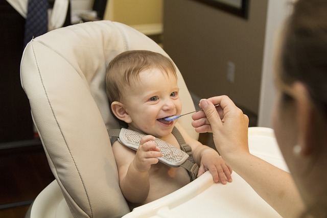 一個微笑的嬰兒坐在高腳椅上，正在由一個成年人用藍色湯匙餵食，背景是溫暖的室內環境。