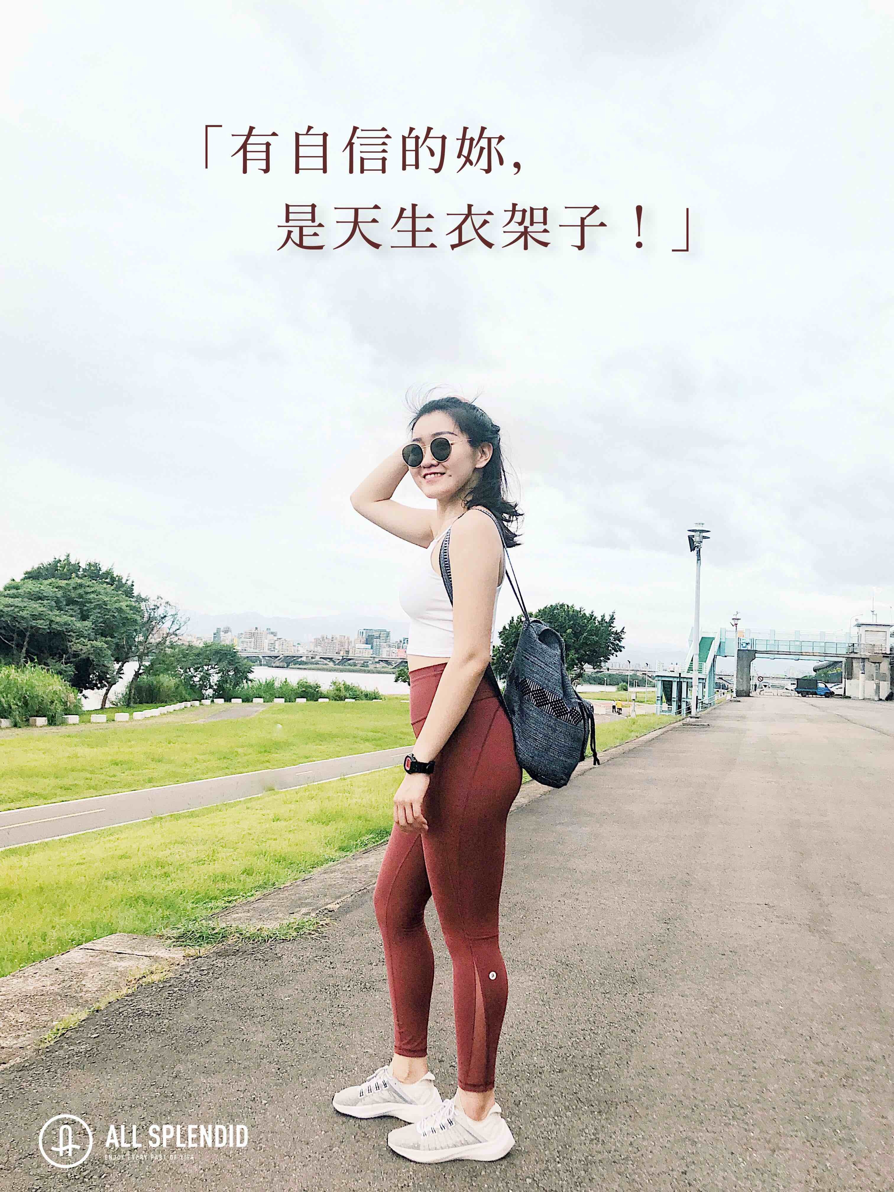 一名年輕女子站在小路上，身穿白背心紅色ASify 瑜伽褲，背著灰色背包。路旁是綠草地，遠景是城市高樓。整體營造出輕鬆自在的生活場景。