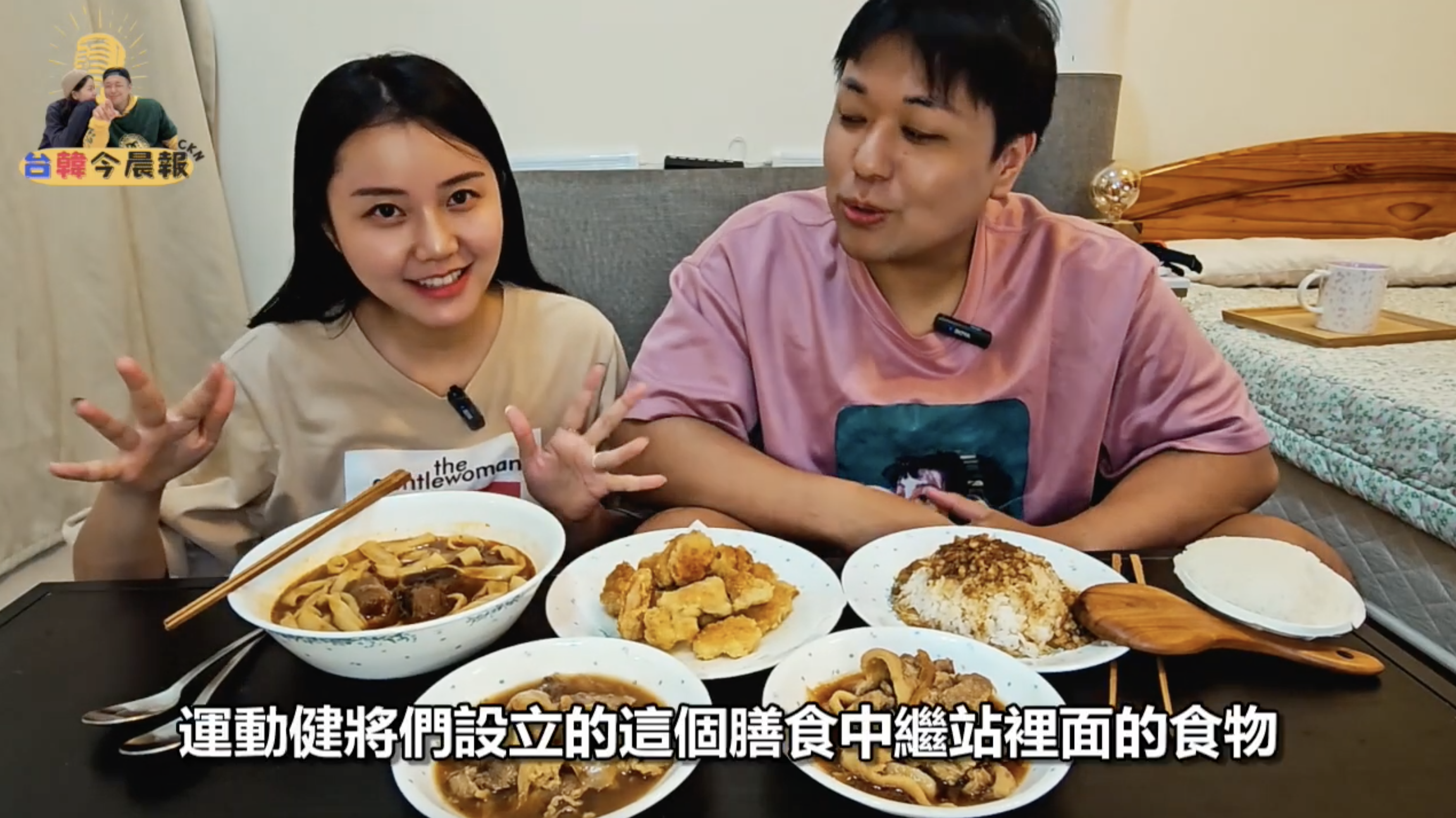 年輕男女坐在桌前,桌上擺放著多道菜餚,包括湯麵、炸物和米飯等。女性正在用手勢解釋,男性則看著食物。畫面下方有字幕"運動健將們設立的這個膳食中繼站裡面的食物"。