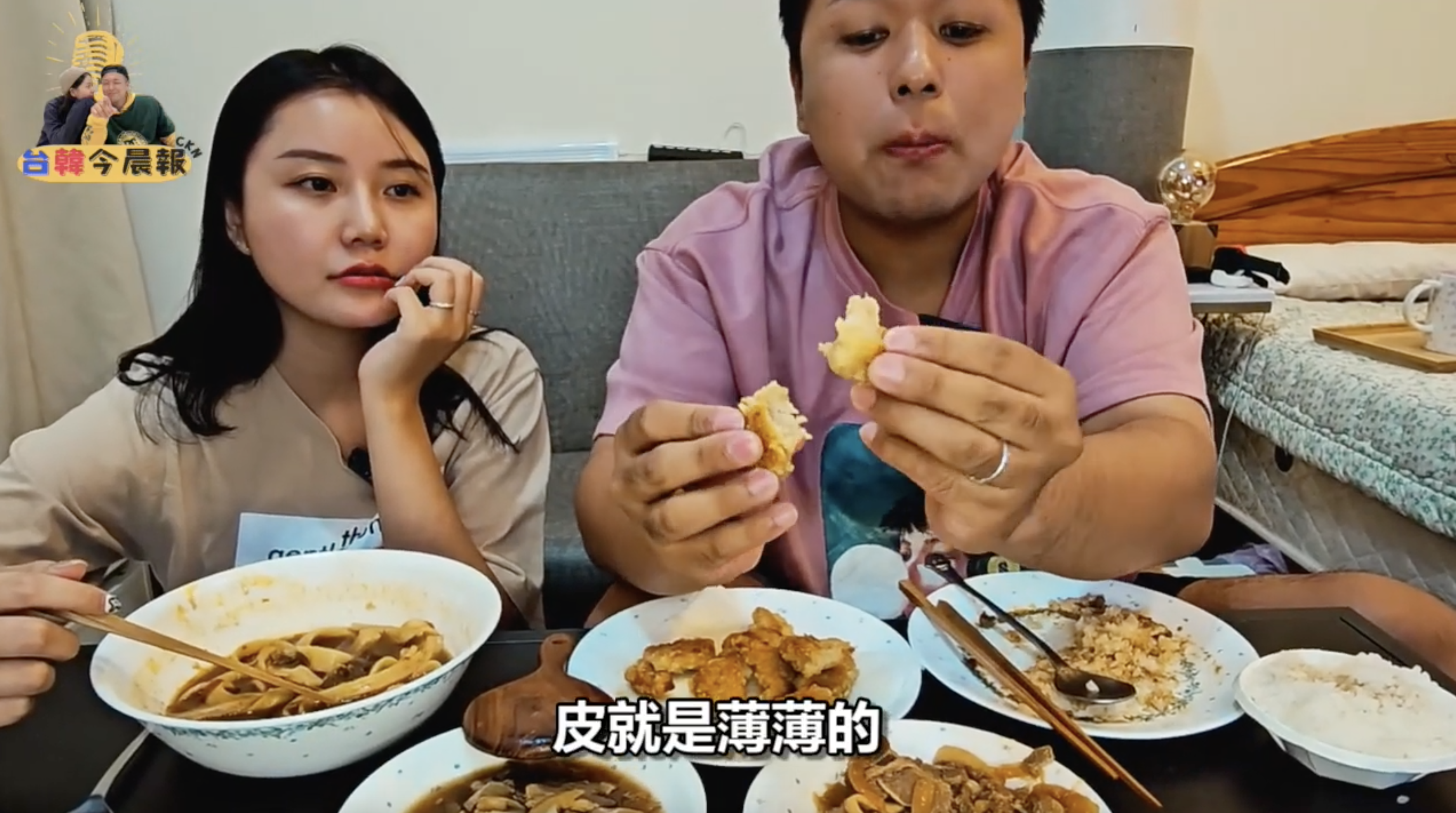 一名年輕女性和一名年輕男性坐在沙發上品嚐多種食物。桌上擺放著湯麵、炸雞和其他菜餚。男性正在品嚐一塊食物，女性則專注地看著。畫面下方有字幕"皮就是薄薄的"。