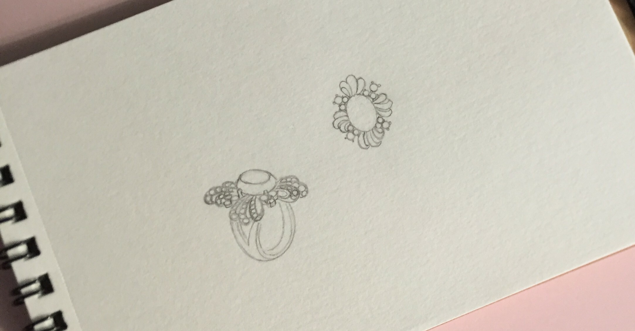A pencil sketch of two jewelry designs features a detailed ring with a central gemstone, surrounded by intricate floral and bead-like elements. Above it, there is a top-down view of a floral-inspired piece with a central stone encircled by petal-like details.