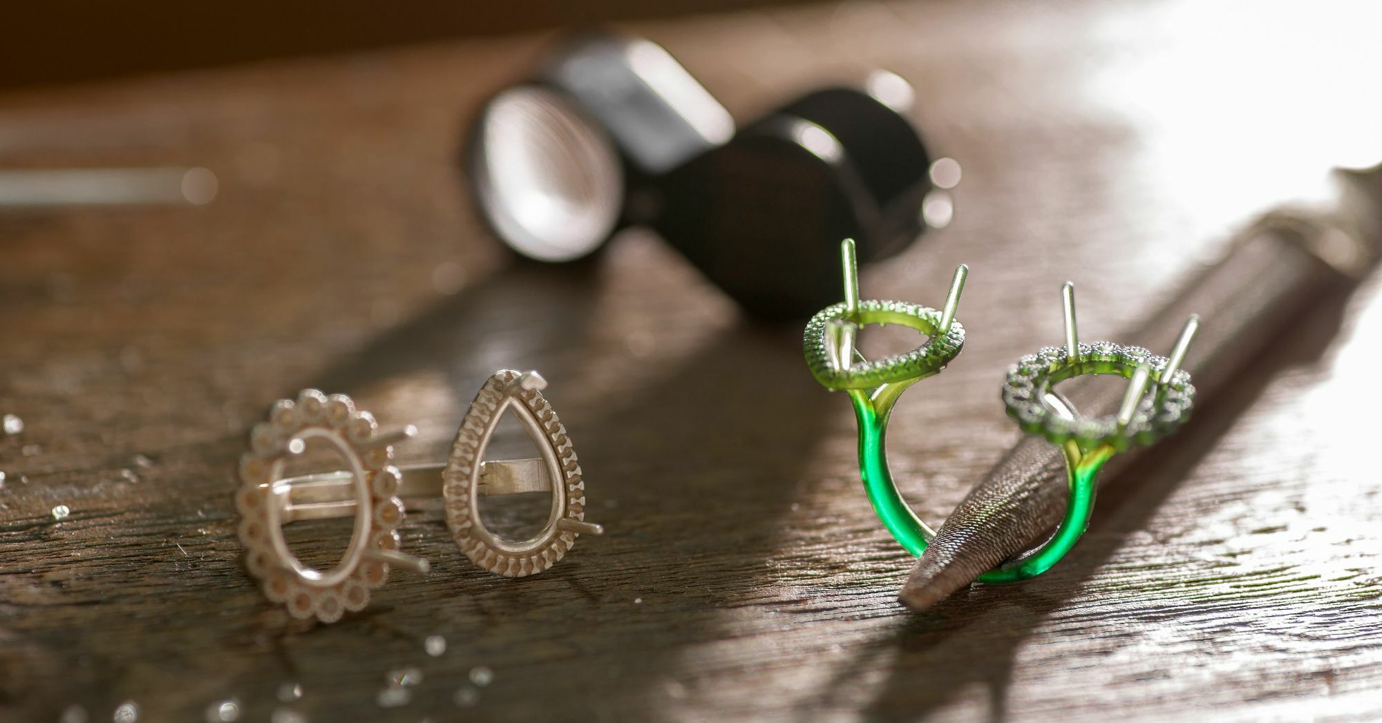 On the left, one silver ring settings features teardrop and floral designs with delicate engraving and small gemstone setting holes. On the right, one green wax ring model displays prongs and intricate settings, ready for the casting process.