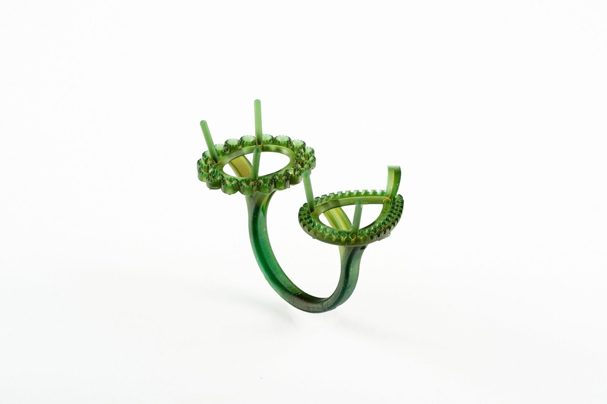 A green wax model of a custom-designed double-ring setting. The ring features two distinct circular settings with intricate prong and pave details, intended to hold gemstones. The band elegantly curves to connect both settings, showcasing a unique and artistic jewelry design before the casting process.