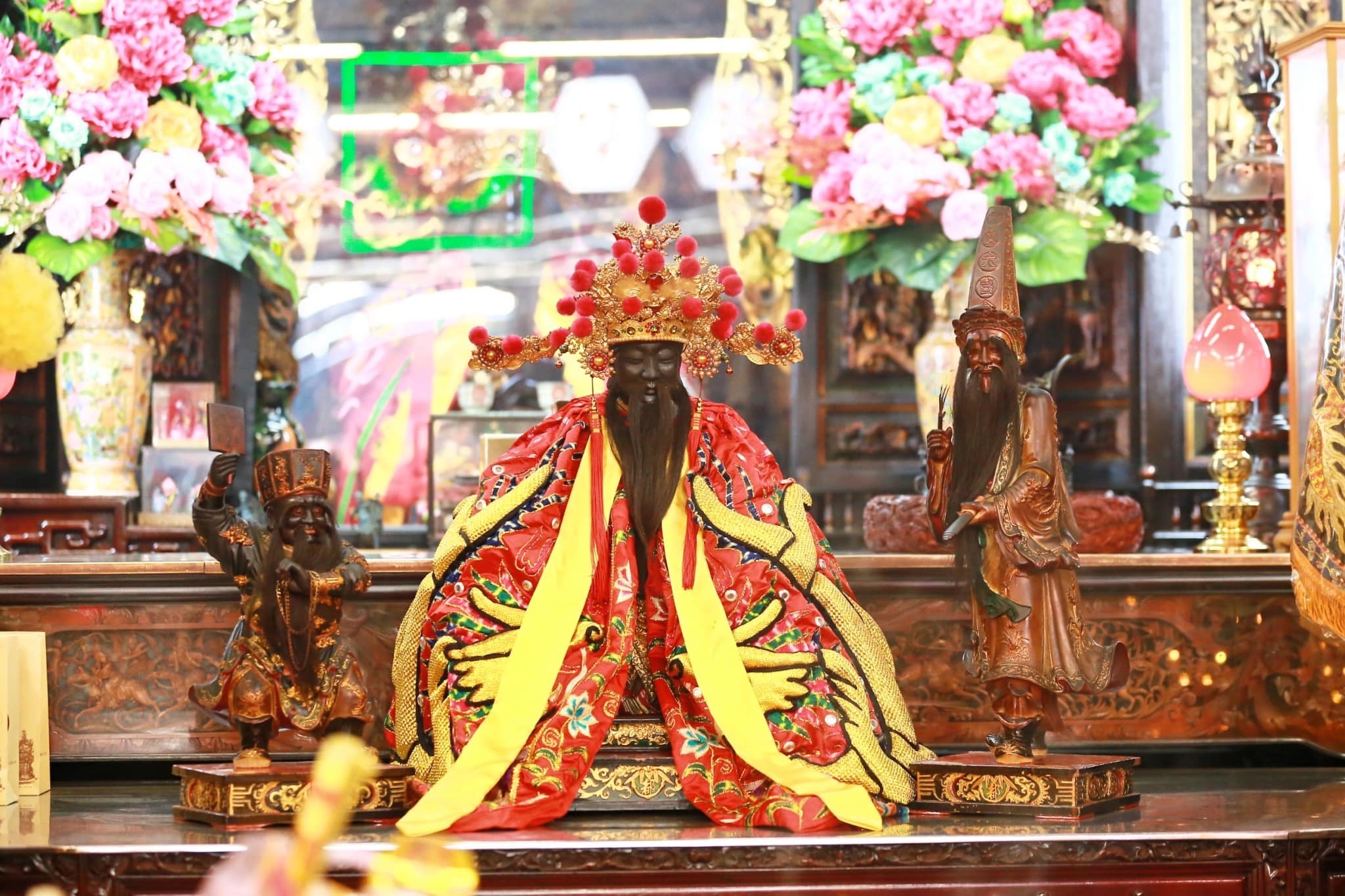 這張圖片顯示的是新竹都城隍爺的神像與大爺二爺。圖中，主神城隍爺身著華麗的紅黃相間的衣袍，頭戴金冠，坐姿威嚴，兩旁分別是大爺和二爺的神像，呈現出一種莊嚴的神聖氛圍。背景裝飾有鮮豔的花卉，突顯出廟宇的神聖與典雅。