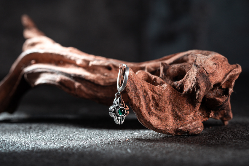 凱爾特之夢 純銀耳墜 Celtic Dream Silver Earrings，以凱爾特盾結為設計主體，中央綠寶石象徵夢境與靈魂守護，吊墜式耳環懸掛於自然木紋背景上，展現神秘靈性與細緻銀飾工藝的結合
