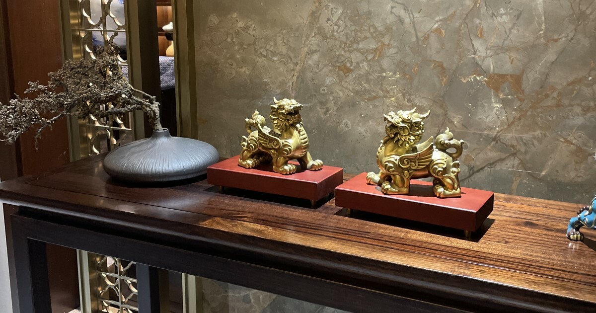金色銅貔貅成對擺放於紅色底座上,設於木製展示檯,背景為中式風格石牆,營造典雅莊重的風水擺設氛圍。