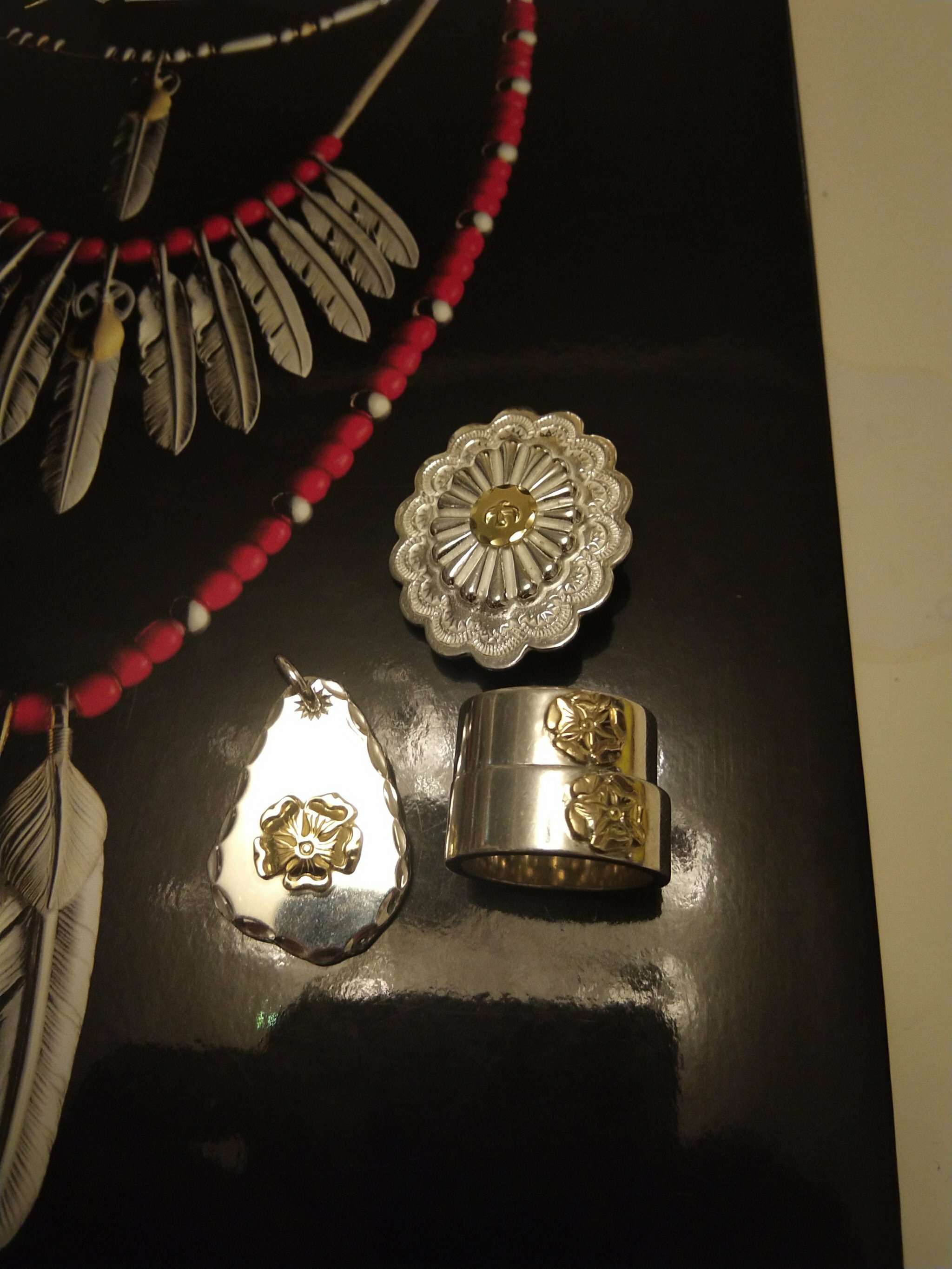 Goro's銀飾配件展示,包含花朵圖騰吊墜、雕花戒指與圓形銀章,背景為羽毛設計海報