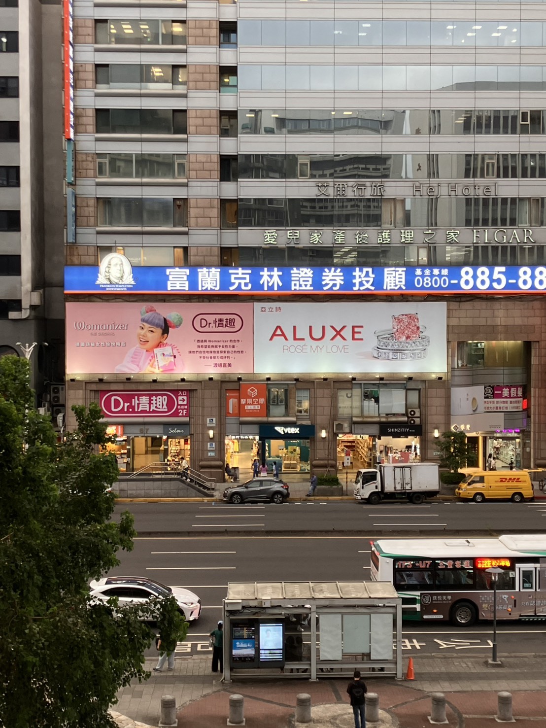 台北市東區忠孝東路上的商業大樓外牆廣告，包括渡邊直美代言的Womanizer與Dr.情趣聯名廣告，底下有Dr.情趣忠孝門市入口與多家商店，街道上有公車站牌與行駛中的車輛。