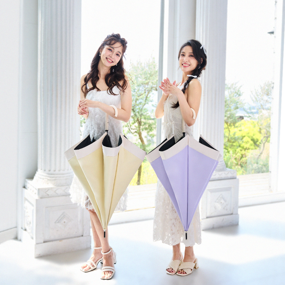 兩位穿著白色洋裝的女性在室內微笑，手中各拿著一把傘，一把為黃色、一把為紫色