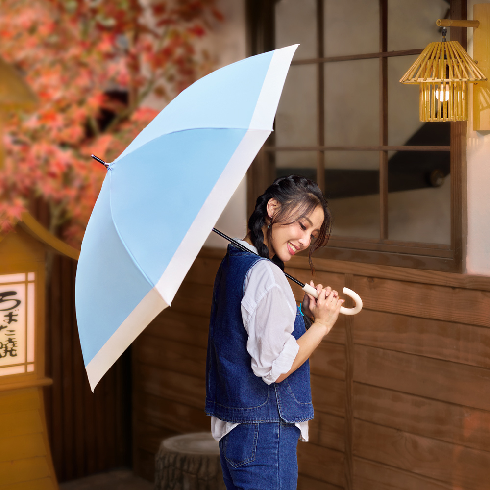 一位年輕女性站在日式風格木屋前，身穿牛仔背心與牛仔褲，手持藍色雨傘微笑