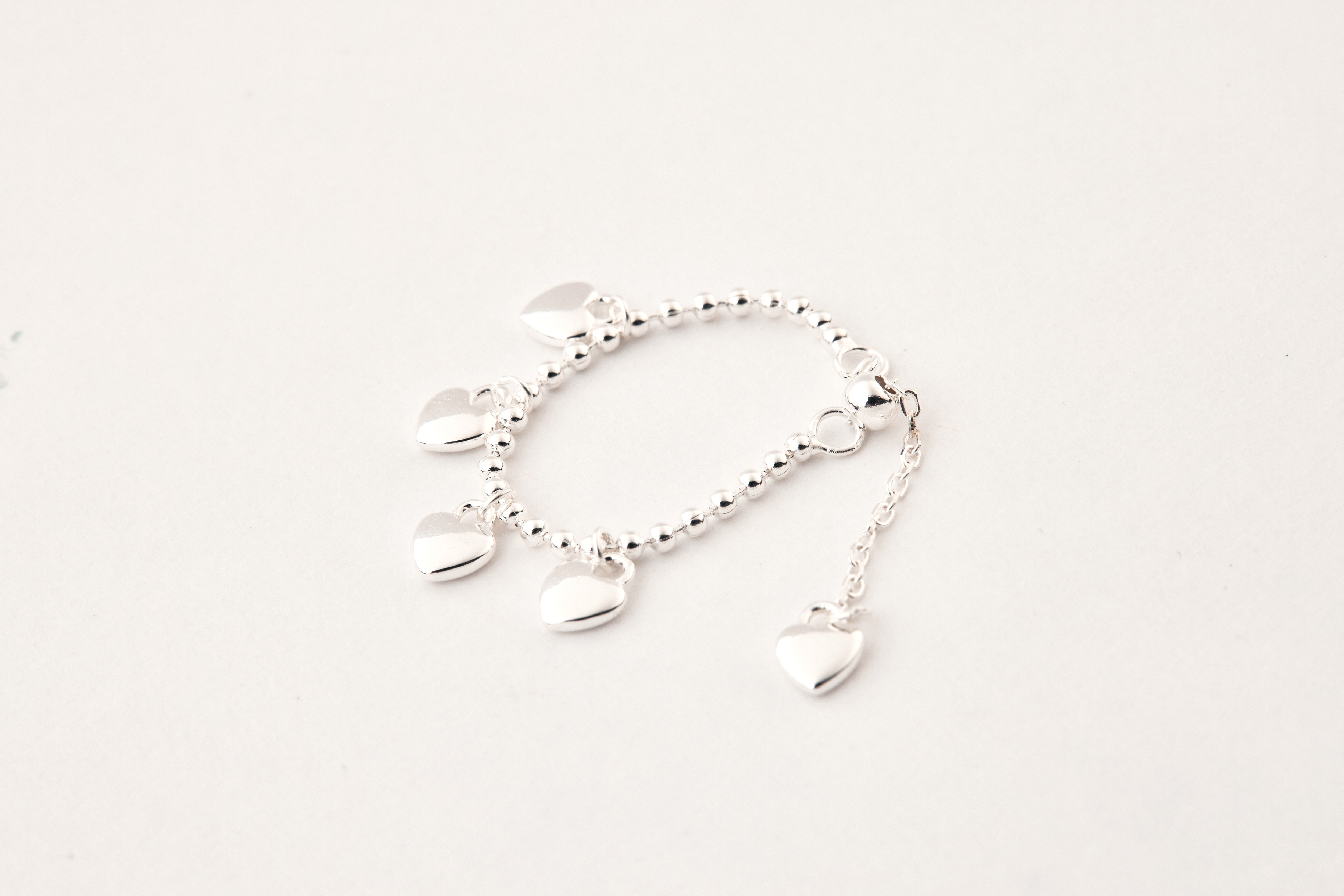 愛心鏈戒平放於淺色背景上，鍊條細緻排列，愛心造型圓潤精緻，整體風格柔和簡約。