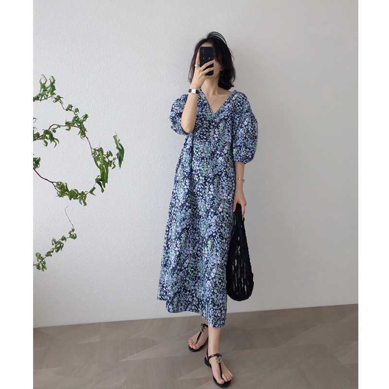 一位女性身穿藍色碎花V領中長連衣裙，搭配黑色編織手提袋和平底涼鞋，在白色牆面前拍攝的夏季穿搭照。