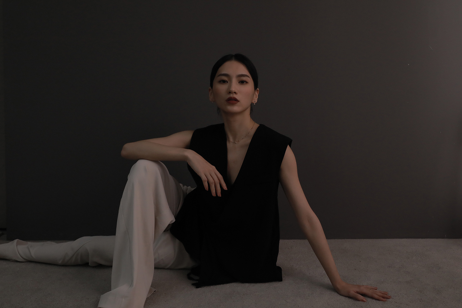 身穿黑色無袖上衣與白色長褲的女性坐於地毯上，背景為深灰色牆面，光影低調柔和，展現沉靜且堅定的優雅氣質。