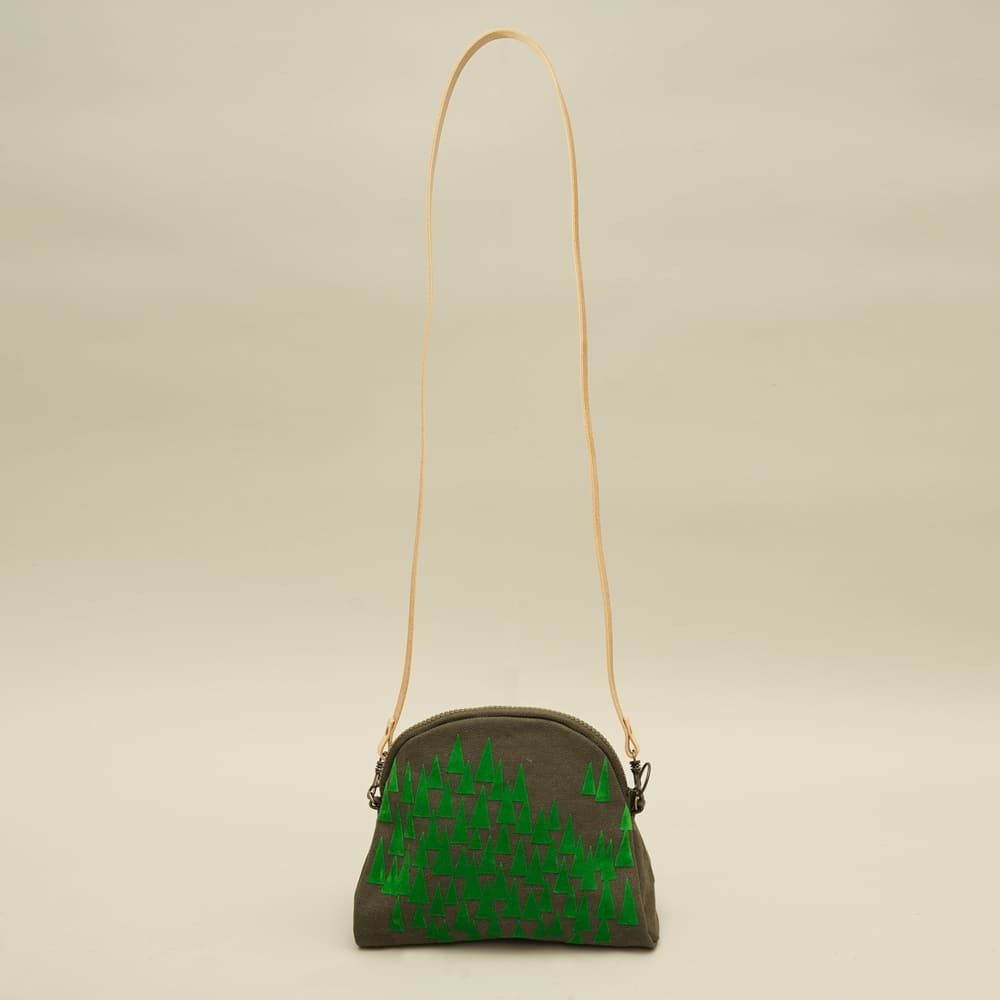 灰綠色帆布斜背包，正面印有多顆綠色三角形松樹圖案，搭配淺色皮革肩帶