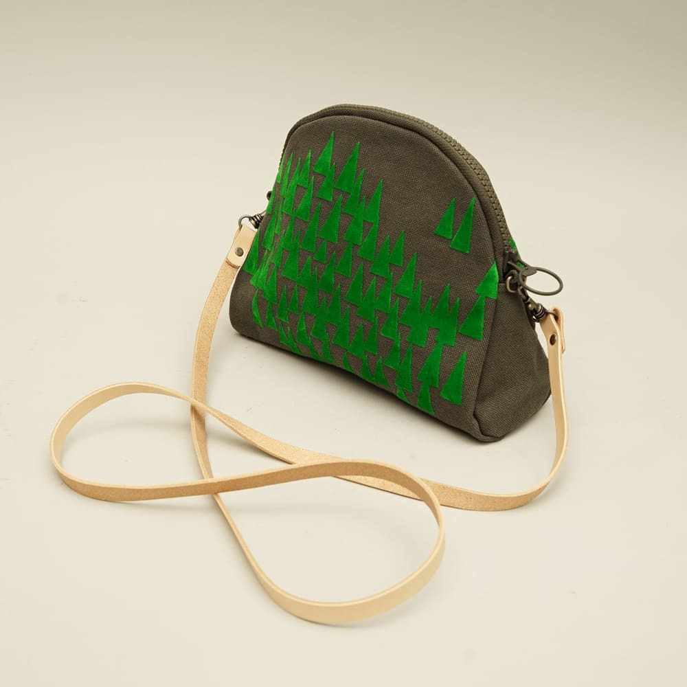 灰綠色帆布斜背包，正面印有多顆綠色三角形松樹圖案，搭配淺色皮革肩帶