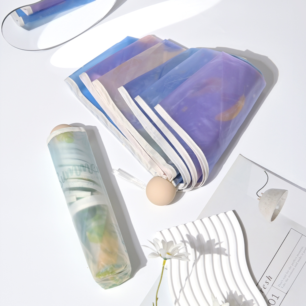 收合的夢幻透明摺疊傘-樹葉款及半打開的夢幻透明摺疊傘-羽毛款放在桌上,擺放著白色小花及筆記本在周圍裝飾。