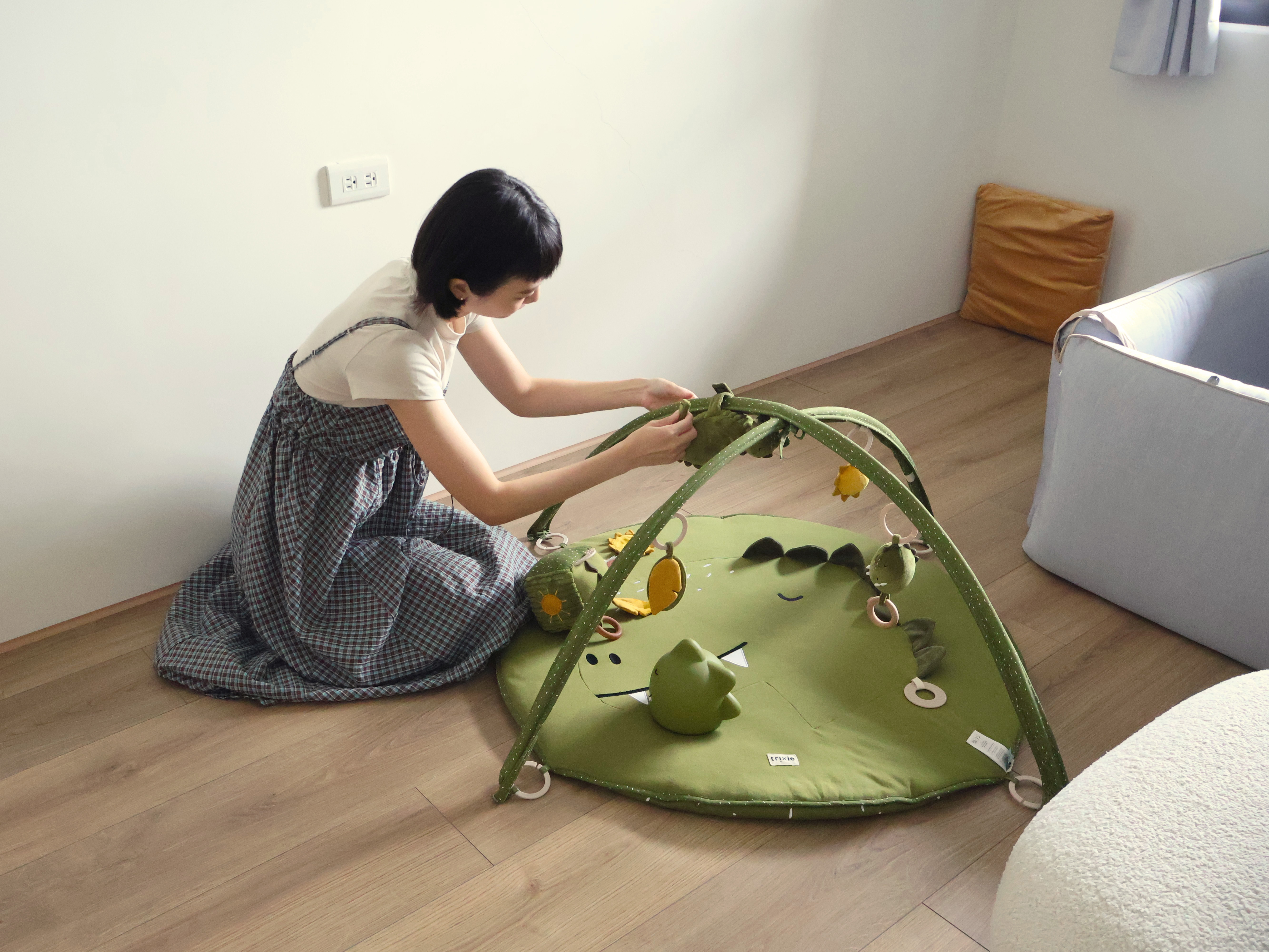 一位穿著格子連身裙的母親跪在木地板上，專注組裝綠色恐龍造型遊戲墊，墊上掛有可愛的動物吊飾，背景有白色牆壁、橙色靠墊和灰色嬰兒圍欄，展現舒適的嬰兒房佈置氛圍。