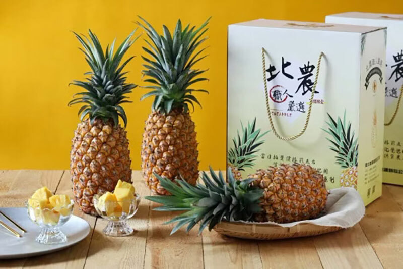 入厝禮盒推薦，三顆新鮮的鳳梨，其中兩顆直立放置於桌面上，另一顆則橫放在淺色的托盤裡。桌面上還擺有兩個小玻璃碟，畫面右側有一個設計精美的紙盒禮袋