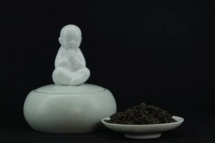 茶席擺設陶瓷小和尚擺件，白色佛像安坐合十於茶桌之上，搭配深色茶具底座，展現茶道中的禪意氛圍，營造寧靜沉穩的品茶環境，常用於茶藝表演與茶室佈置，象徵靜心與專注。