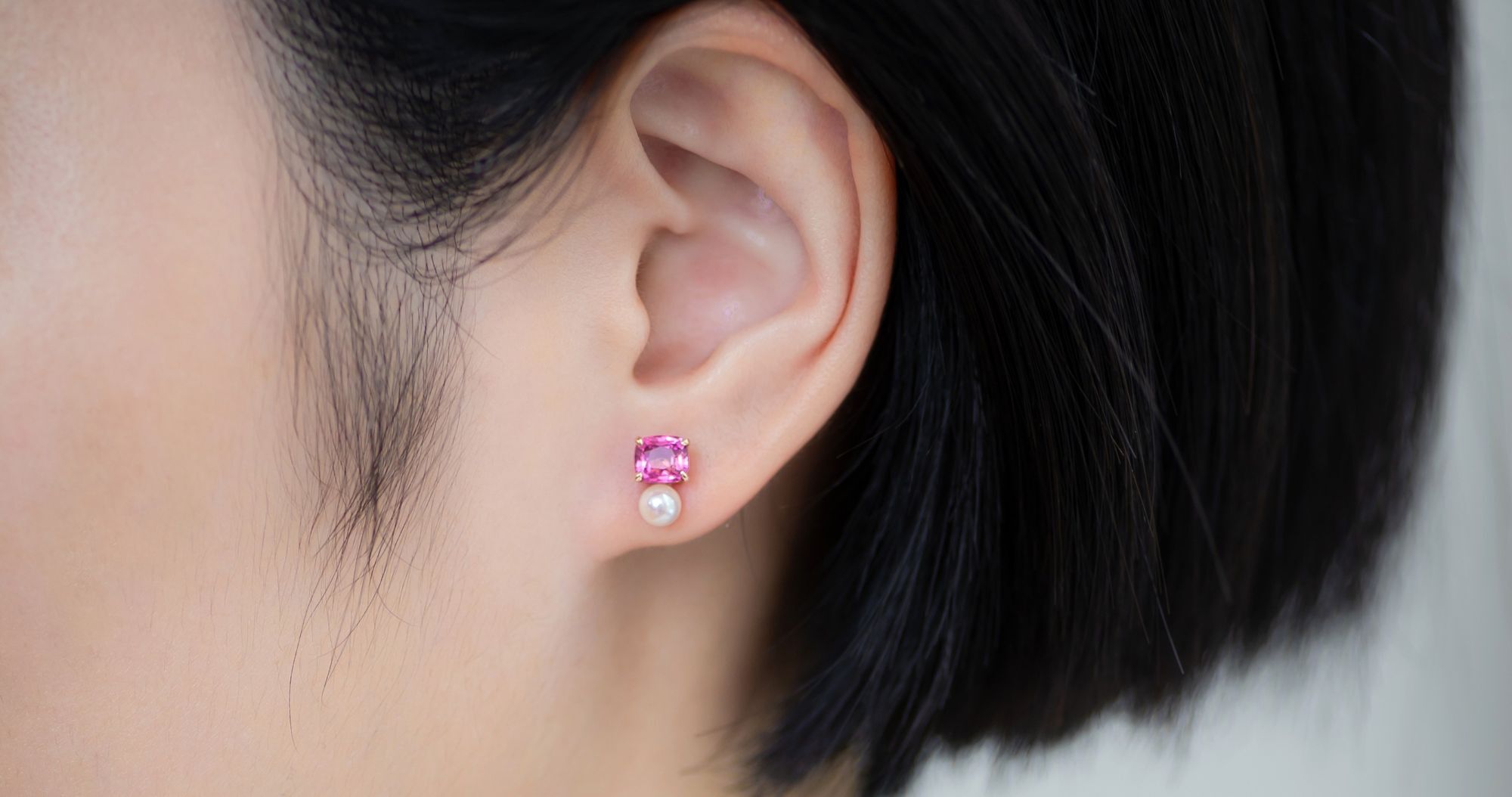 A woman's ear wearing a delicate earring featuring a vivid pink spinel paired with a small, round Akoya pearl, set in a minimalist design.