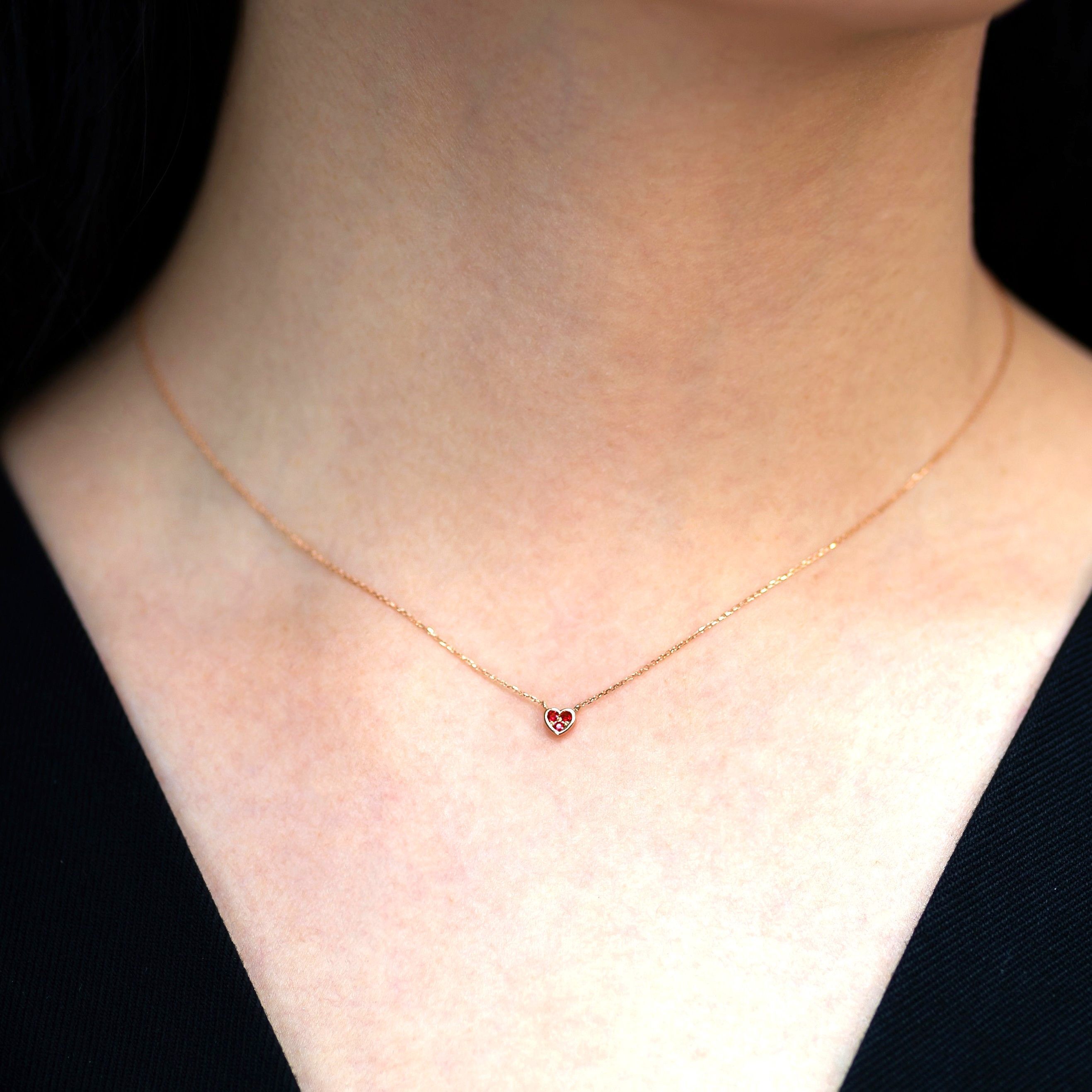 Close-up of a woman's neck wearing a delicate gold necklace with a small red heart-shaped ruby pendant.