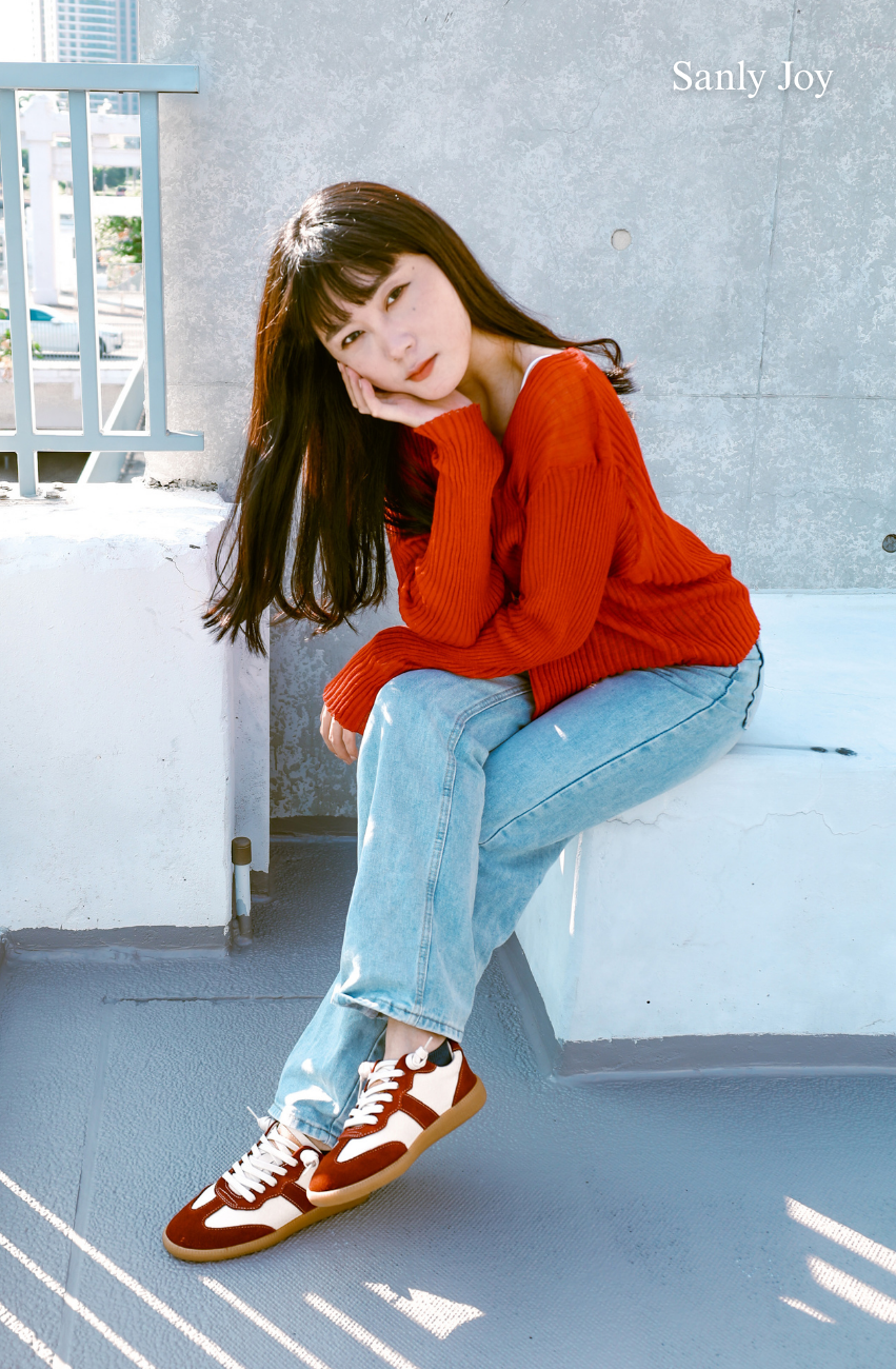 Sanly Joy 蘋果牛奶真皮德訓鞋形象照,模特著紅色針織上衣與淺色牛仔褲,腳穿紅白拼接真皮德訓鞋,展現秋冬自然率性的休閒氛圍。鞋款以柔軟牛皮與麂皮拼接打造,鞋底輕盈舒適,行走穩定不咬腳,呈現兼具質感與舒適度的日常穿搭。