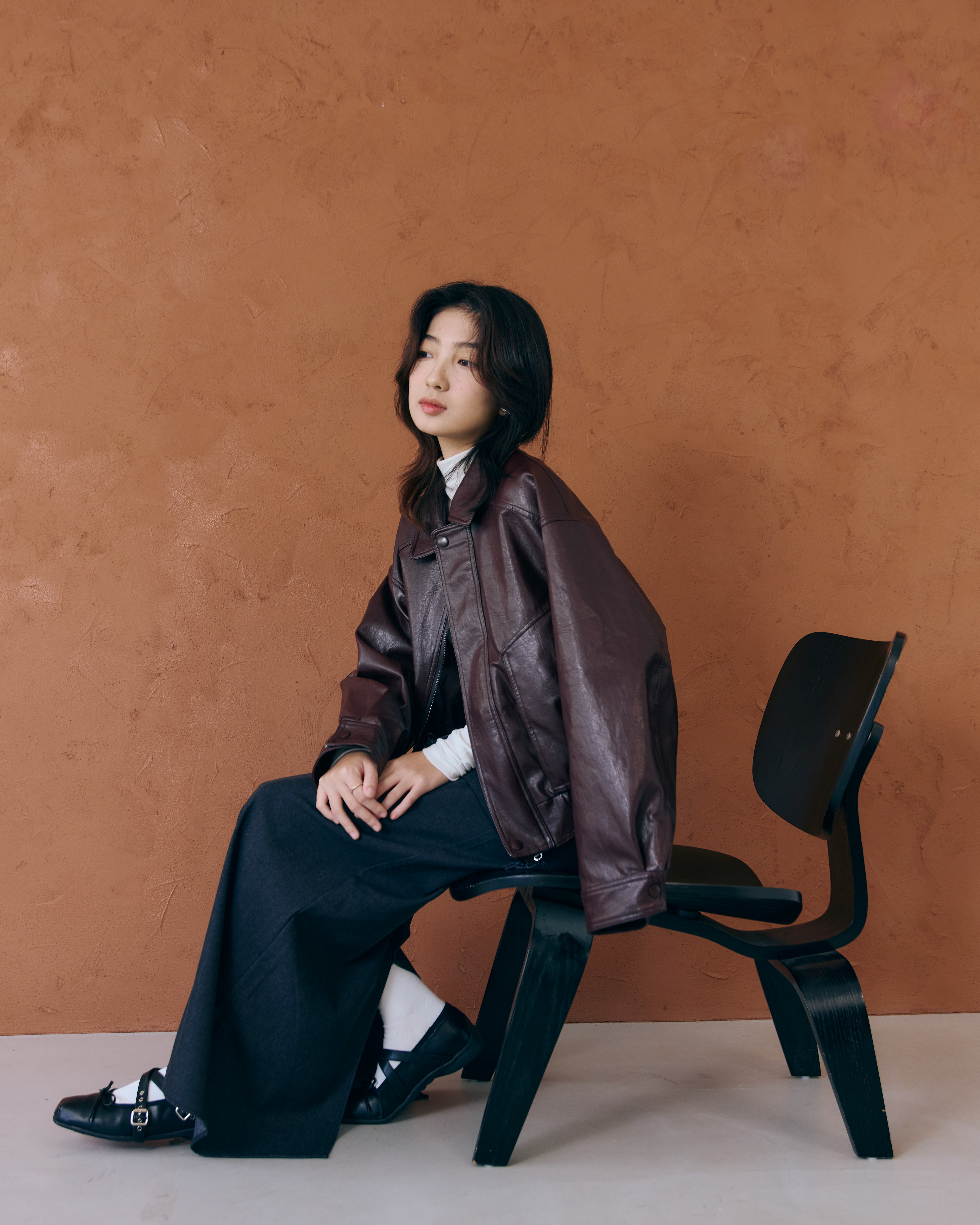 一個女生穿著版型俐落帥氣的酒紅皮衣，下身穿直筒裙坐在椅子上，雖然穿著帥氣的皮衣但配裙子整體給人一種女人味高雅感