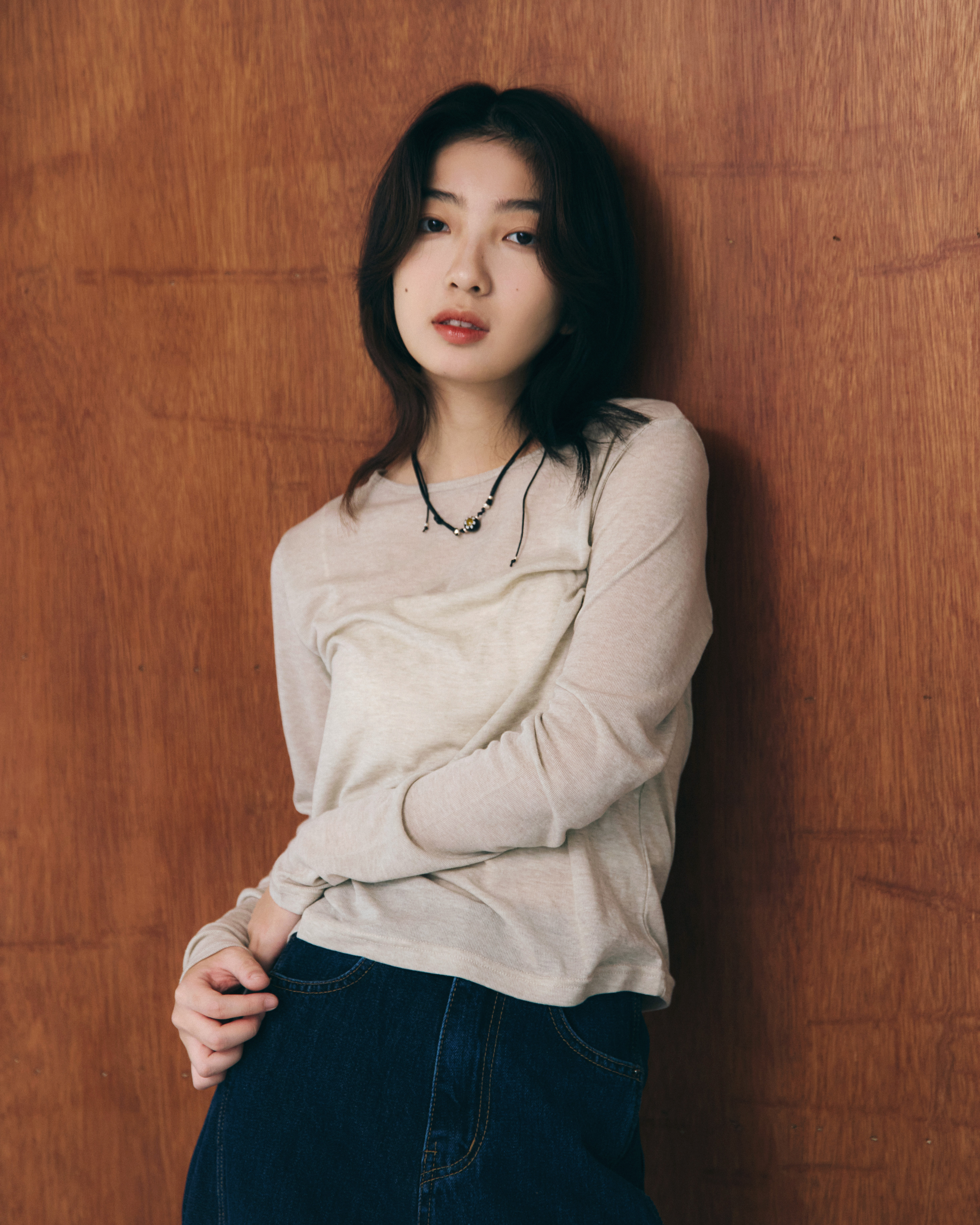 一個女孩靠在牆上，衣服穿著摩卡色的薄長袖，並且用有墜飾的項鍊點綴整體穿搭