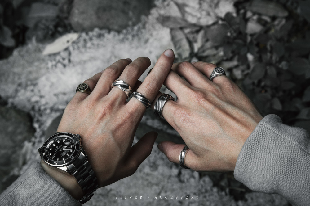 近景雙手配戴多枚純銀戒指與金屬腕錶的穿搭示意，冷調石面背景；主打款為 JK銀飾「鋼鐵之韌」純銀戒指，呈現寬版層次與金屬光澤，適合男士銀飾疊戴造型。