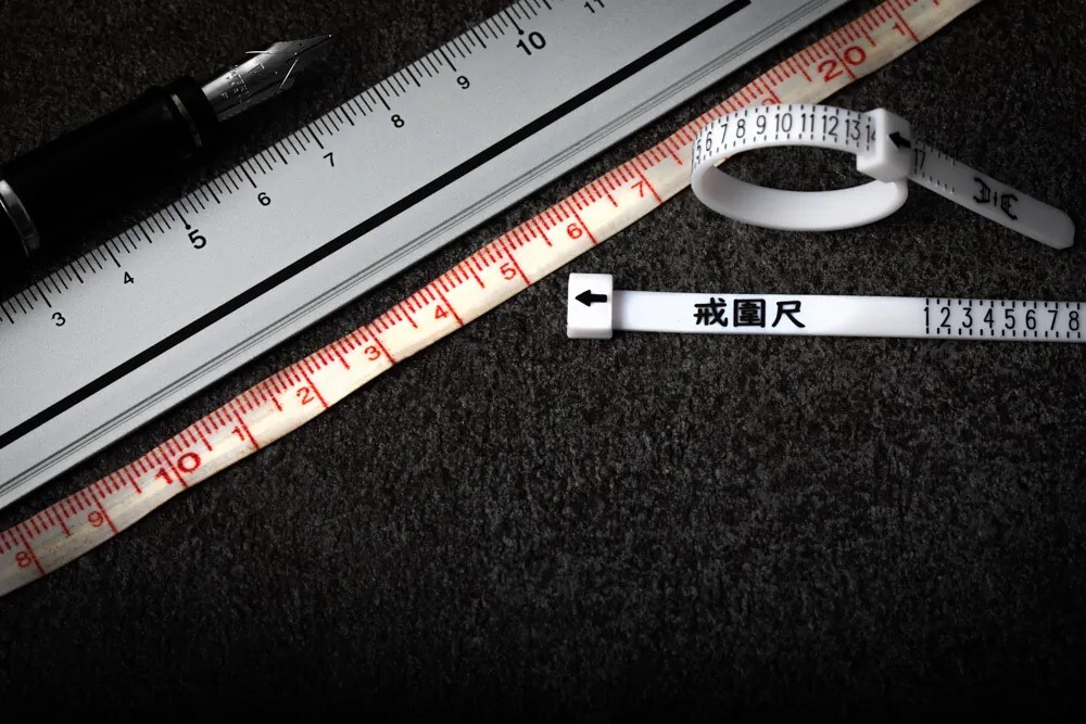 桌面近拍：直尺、皮尺與可調式「戒圍尺」（1–13 號）量測工具擺放示意，用於戒指尺寸測量與內徑/周長換算；網購前自助量戒圍指南（JK銀飾）。