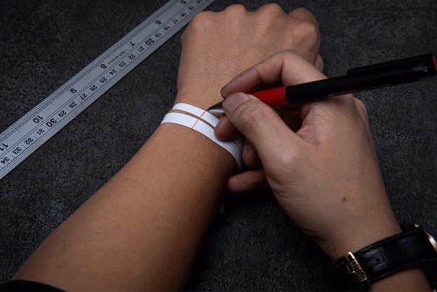 手圍測量示意：在腕骨上方一指處以無彈性紙條貼合繞腕，交會點以筆做記號；旁置毫米直尺，攤平後量長取得手圍，用於手鍊／手環尺寸選擇。