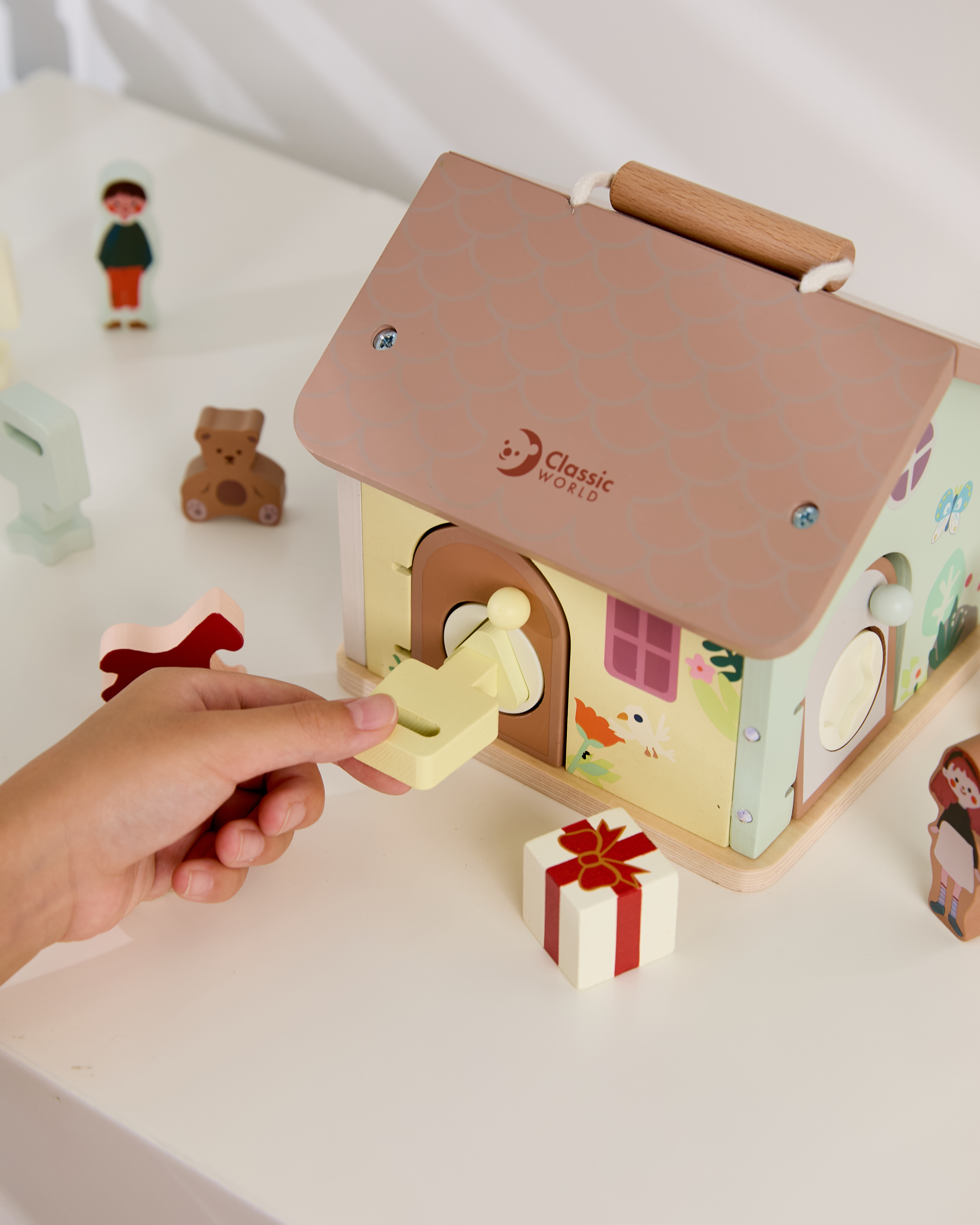 一個孩子的手正拿著一把淡黃色的鑰匙，試圖打開 Classic World 開鎖小木屋 這款木質感統玩具上的門鎖。小木屋有著粉棕色的屋頂，側邊有精美的圖案裝飾。周圍散落著其他木製配件，如小熊、人物剪影和一個小禮物盒。