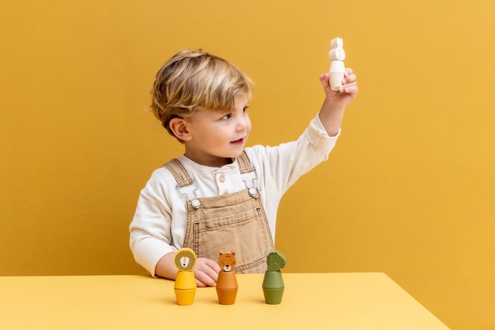 一位幼兒（約兩歲）坐在黃色桌前，背景為亮黃色，他正使用一隻手將兩個動物造型的木製零件（可能是堆疊或螺絲結構）組合起來，並高舉觀察。桌面上擺放著三個可愛的動物造型底座，分別是黃色的獅子、棕色的老虎和綠色的恐龍。