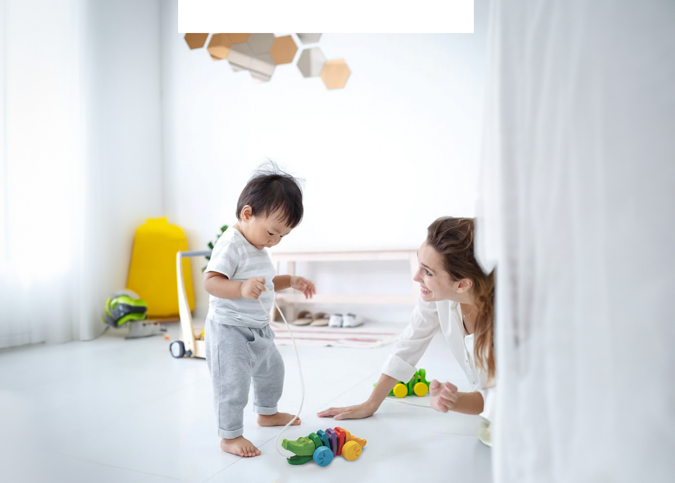 一位穿著白色上衣和灰色長褲的幼兒，光著腳站在明亮的室內地板上，正俯身準備拉動一個色彩鮮豔的 Plantoys 彩虹鱷魚拉車。一位成年女性面帶微笑地在旁邊看顧與引導。 (H3) Plantoys 彩虹鱷魚拉車｜邁開第一步的節奏夥伴