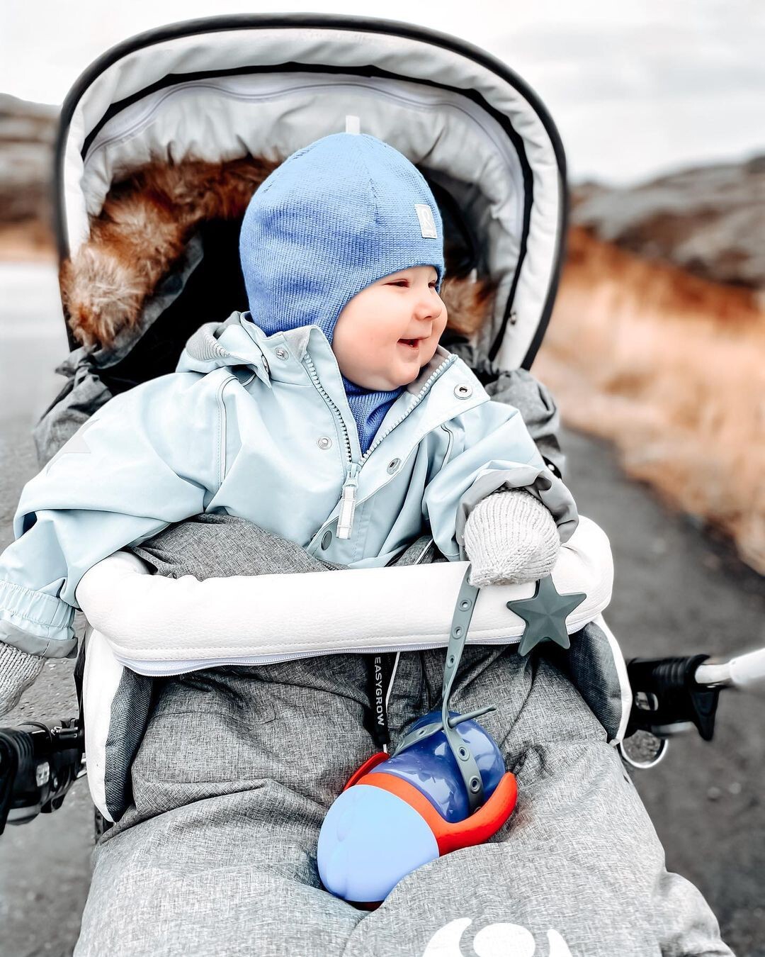 一位穿著藍色外套的寶寶坐在嬰兒車中，一個灰色的固齒防掉帶將他的水壺固定在嬰兒車扶手上。