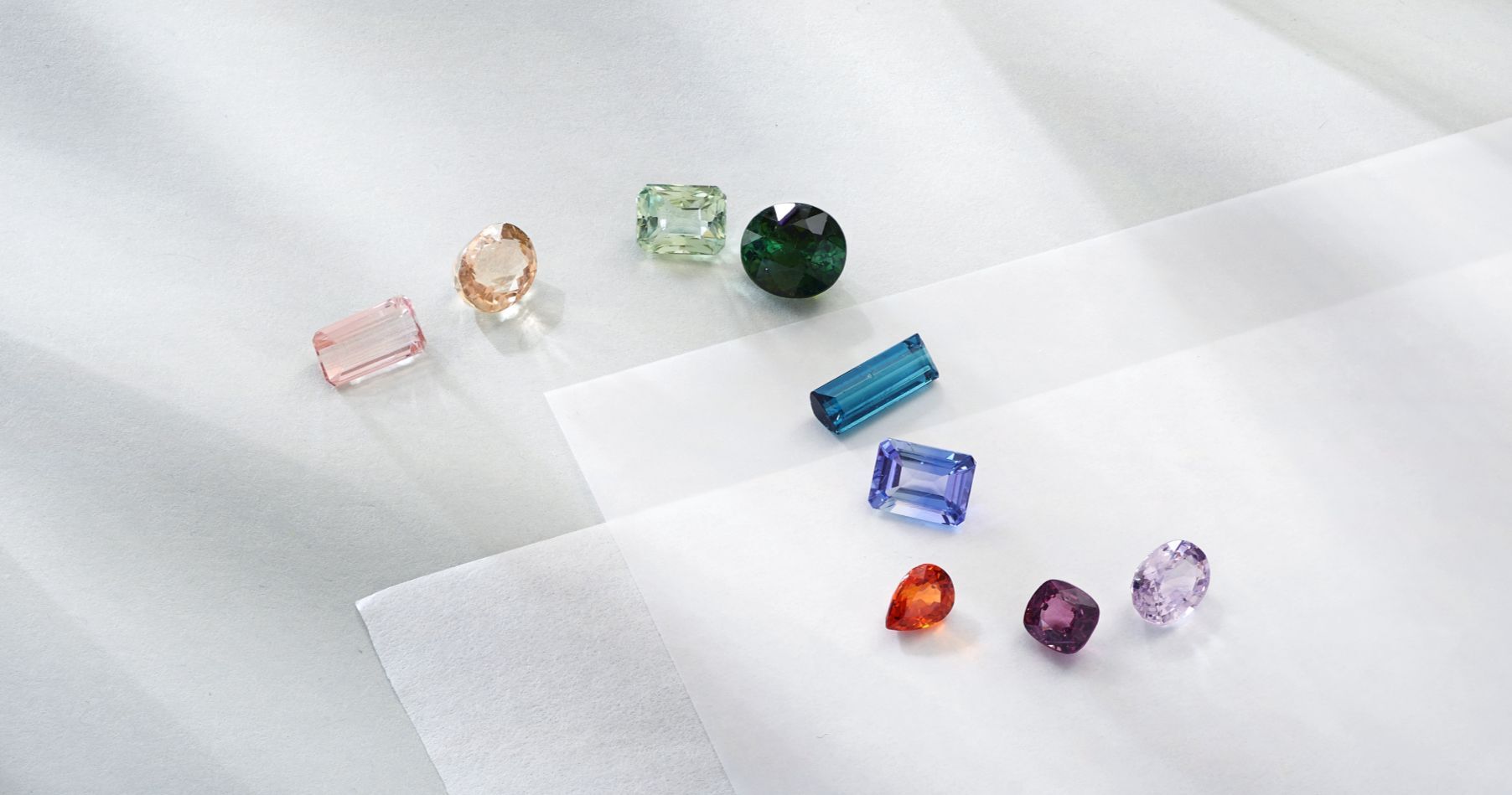 A collection of colorful loose gemstones in various shapes and cuts, including pink, peach, green, dark green, blue, purple, orange, and lavender, displayed on soft white and light grey textured paper.