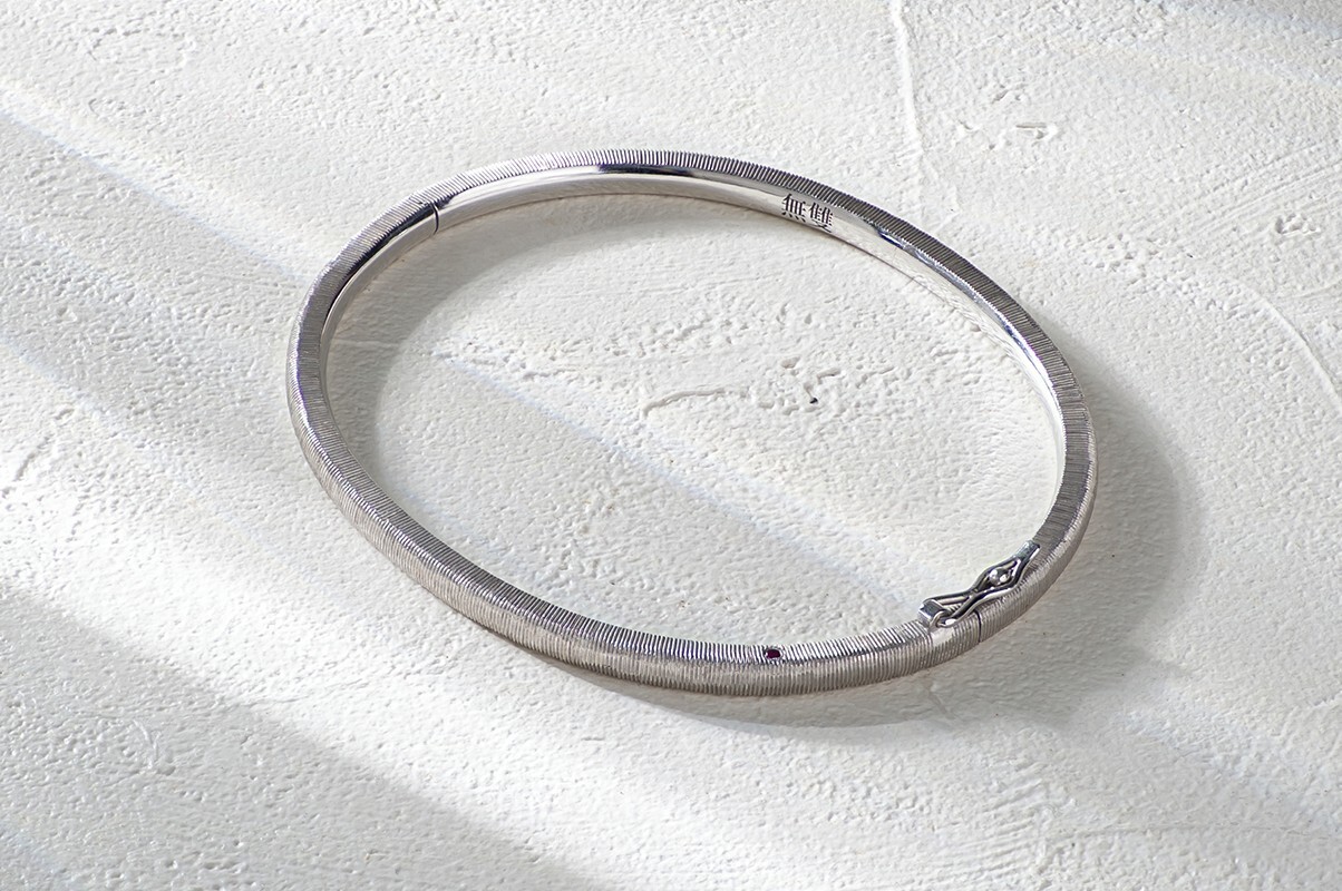 A bespoke S925 silver and palladium alloy bangle with a carved, brushed texture, resting on a textured white surface in sunlight.