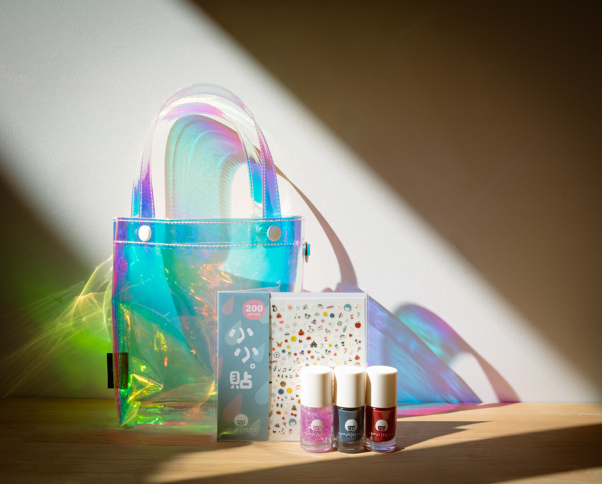 「小小PETIT × 理理 liil 冬夜花火聯名小小微光包｜透明虹光托特包搭配三款無毒水性可撕指甲油與小小貼貼，置於木質桌面與自然光影下的限定商品陳列照」